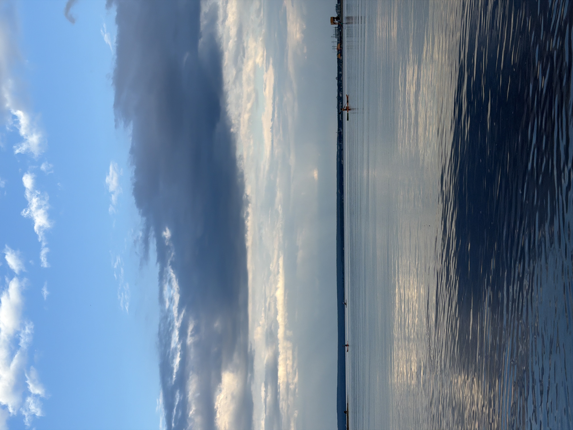 The image depicts a wide, calm body of water, likely a lake or a large river, in Noviy, Russia. The sky above is a mixture of bright blue and dramatic, dark clouds, with shafts of sunlight piercing through the latter. The water's surface is mostly still, reflecting the sky and distant shoreline.
On the water, three individuals are visible, each in a kayak. One kayak, occupied by a person in an orange life vest, is positioned towards the right side of the frame, further from the viewer. To the left of this kayak, and closer to the center of the image, is another kayak with an individual. Further to the left, near the distant shore, a third kayak with a person is visible, along with a faint silhouette of a person on the shore.
The distant shoreline appears low and is lined with trees, with some structures and what appear to be industrial elements, possibly a port or power plant, visible on the right side. The overall impression is one of a serene, yet somewhat dramatic, natural landscape.