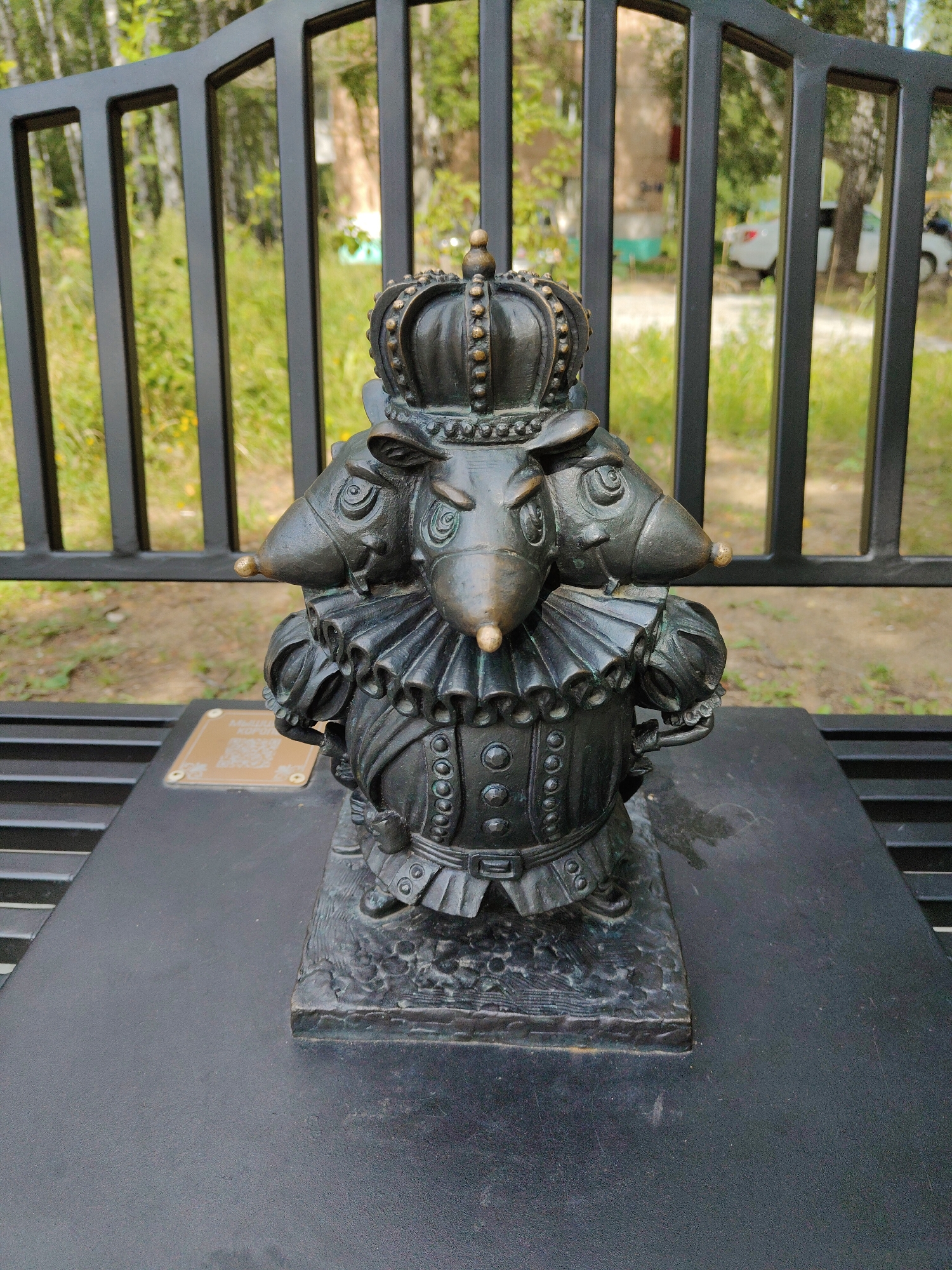 A bronze statue of a multi-headed mouse king stands on a black bench in a park. The statue depicts a mouse with three heads, adorned with a crown and a ruffled collar, wearing a regal outfit with buttons and a belt. The mouse king's heads are facing different directions, with golden-tipped noses. The statue is placed on a dark, textured base, which sits on the black slats of the bench. Behind the bench, a black metal fence separates the park area from a grassy background with trees and a hint of buildings and a white car in the distance. A small golden plaque with a QR code is visible on the bench in front of the statue. The lighting suggests it is daytime.