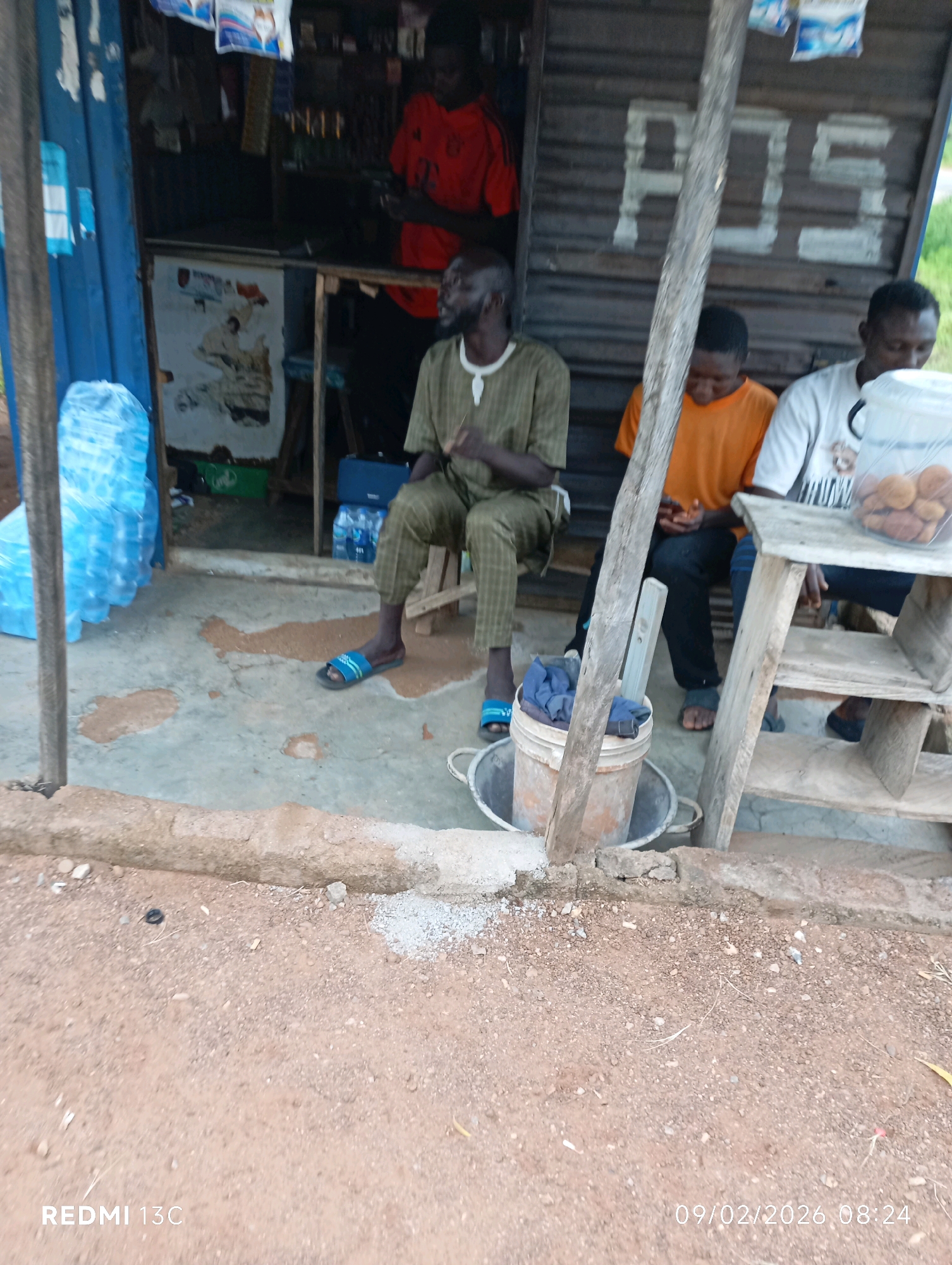In Shendam, Nigeria, three individuals are present near a small roadside stall. The stall has a weathered wooden facade with "POS" painted on it. Two individuals are seated on a wooden structure, and another is seated on a stool. One man is wearing a green and brown patterned shirt and pants, while another wears an orange t-shirt and dark pants. The third man is wearing a white t-shirt with a graphic design. A bucket containing blue cloth is placed on the ground in front of the seated individuals. To the left of the stall, several large plastic bottles of water are stacked. The ground in front of the stall appears to be a dirt and concrete surface. In the background, a person in a red shirt is visible inside the stall. The overall scene depicts a casual gathering at a small business establishment.