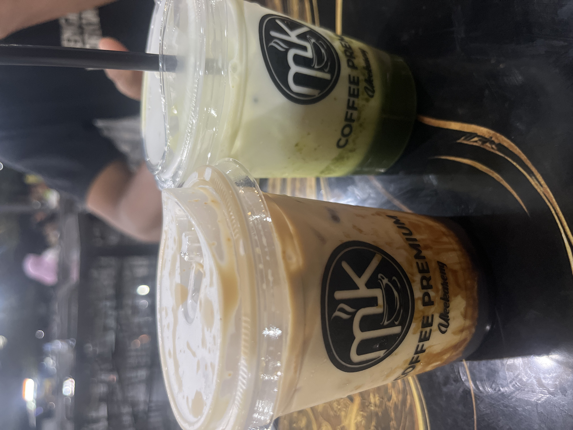 The image depicts two iced beverages in clear plastic cups positioned on a dark, reflective table with swirling gold patterns. The scene is located in Glameunasah Baro, Indonesia.
The cup on the left contains an iced coffee, characterized by a light brown liquid with visible ice and dark caramel or syrup streaks at the bottom. It is sealed with a clear plastic dome lid, which exhibits condensation and small liquid droplets.
The cup on the right contains a layered iced beverage, likely a matcha or green tea latte, displaying a dark green layer at the bottom, transitioning to a lighter green, and topped with a substantial white layer of milk or cream. A black drinking straw is inserted through its clear plastic dome lid, which also shows condensation.
Both cups feature an identical circular black label with white text. The primary logo is "mk" styled with a coffee cup icon beneath the 'm'. Below this, the text "COFFEE PREMIUM" is displayed, with "Ulee Kareng" written in a script font underneath.
The background is heavily blurred, indicating an indoor setting, likely a cafe or restaurant. Blurred figures are discernible; one on the far left wears a light pink head covering. A person's arm, clad in a dark short-sleeved garment with faint white text, is visible on the right side of the frame, positioned near the right beverage. Numerous diffused light sources are present in the blurred background.