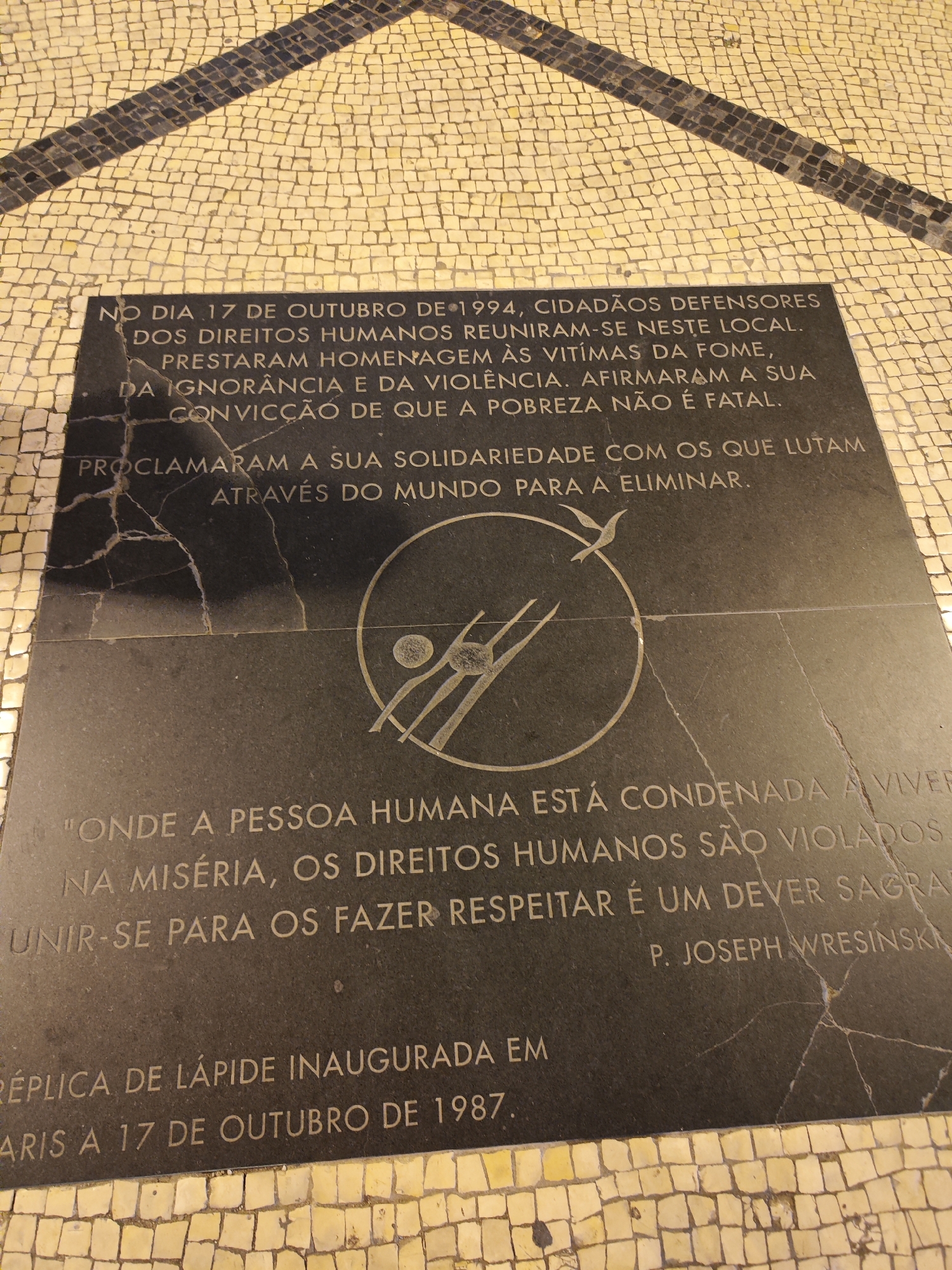 A stone plaque, cracked in several places, is inlaid into a cobblestone walkway. The plaque is inscribed with Portuguese text, which includes the phrase, “NO DIA 17 DE OUTUBRO DE 1994, CIDADÃOS DEFENSORES DOS DIREITOS HUMANOS REUNIRAM-SE NESTE LOCAL.” A circular emblem with a stylized drawing of human figures and a bird is centered on the plaque. The inscription also includes a quote attributed to P. Joseph Wresinski. Text at the bottom of the plaque states it is a replica of a stone inaugurated in Paris on October 17, 1987. The cobblestone walkway surrounding the plaque is patterned in light yellow and black hues.