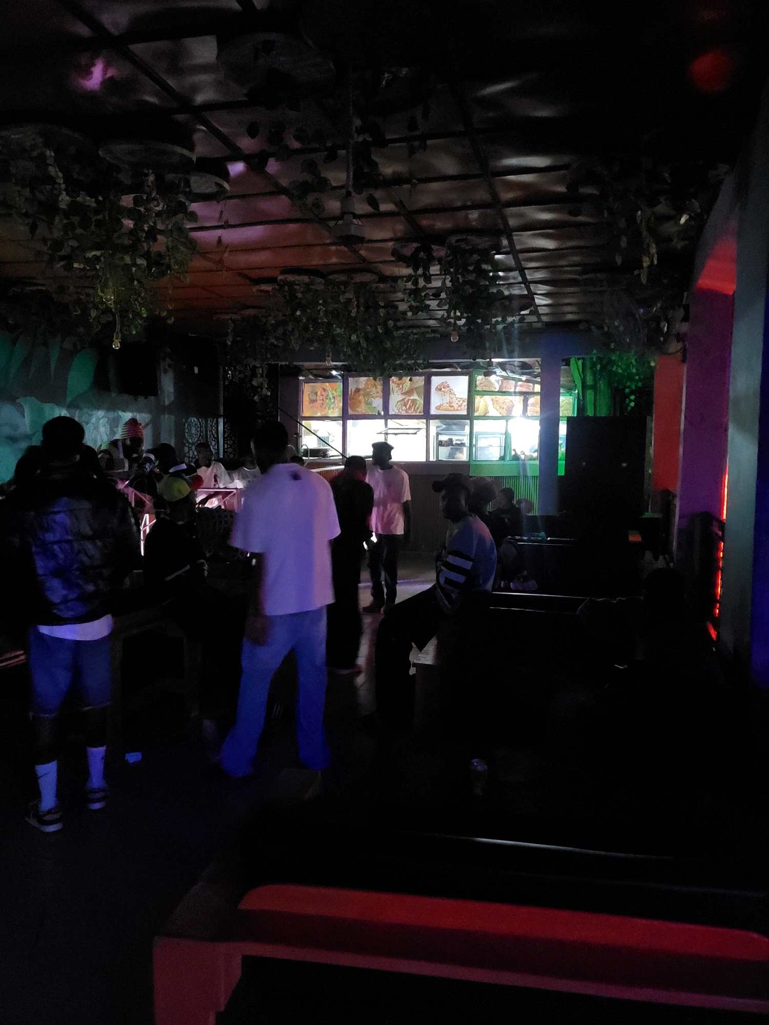 The image depicts an indoor scene, likely a dimly lit establishment, possibly a restaurant or club, in Jos, Nigeria. The ceiling is adorned with hanging artificial foliage and appears to be constructed of metal beams. In the background, a food display with illuminated pictures of pizzas and other dishes is visible through glass panels. Several individuals are present, mostly appearing to be young adults, engaged in various activities. Some are standing, others are seated, and their interactions are not clearly discernible due to the low light conditions and the distance from the camera. The overall ambiance suggests a casual gathering.