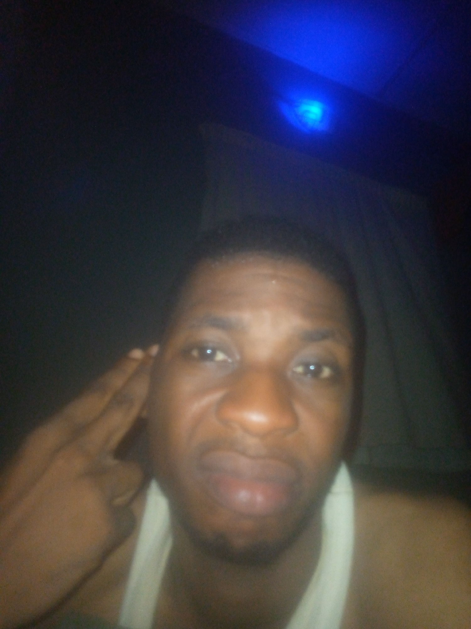 A medium close-up selfie photograph shows a dark-skinned man with short, dark hair, wearing a white tank top. His hand is raised near his head. The location is unknown, though the background suggests an indoor setting with a curtain or sheet. A blue light fixture is visible in the ceiling. The lighting is dim.