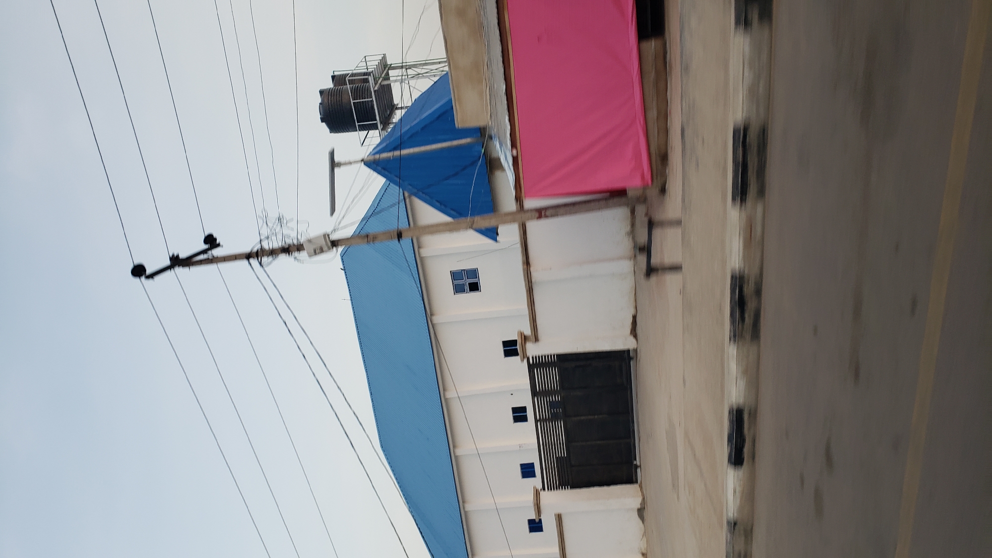 The image shows a white building with a blue metal roof in Jalingo, Nigeria. There is a black metal gate in front of the building, and a white fence. To the right of the building, there is a blue triangular roof structure and a water tower with two black tanks. A pink tarp is draped over something to the right of the gate. Power lines and a utility pole are visible in the foreground. The scene appears to be a street view, with a road in the lower portion of the image.