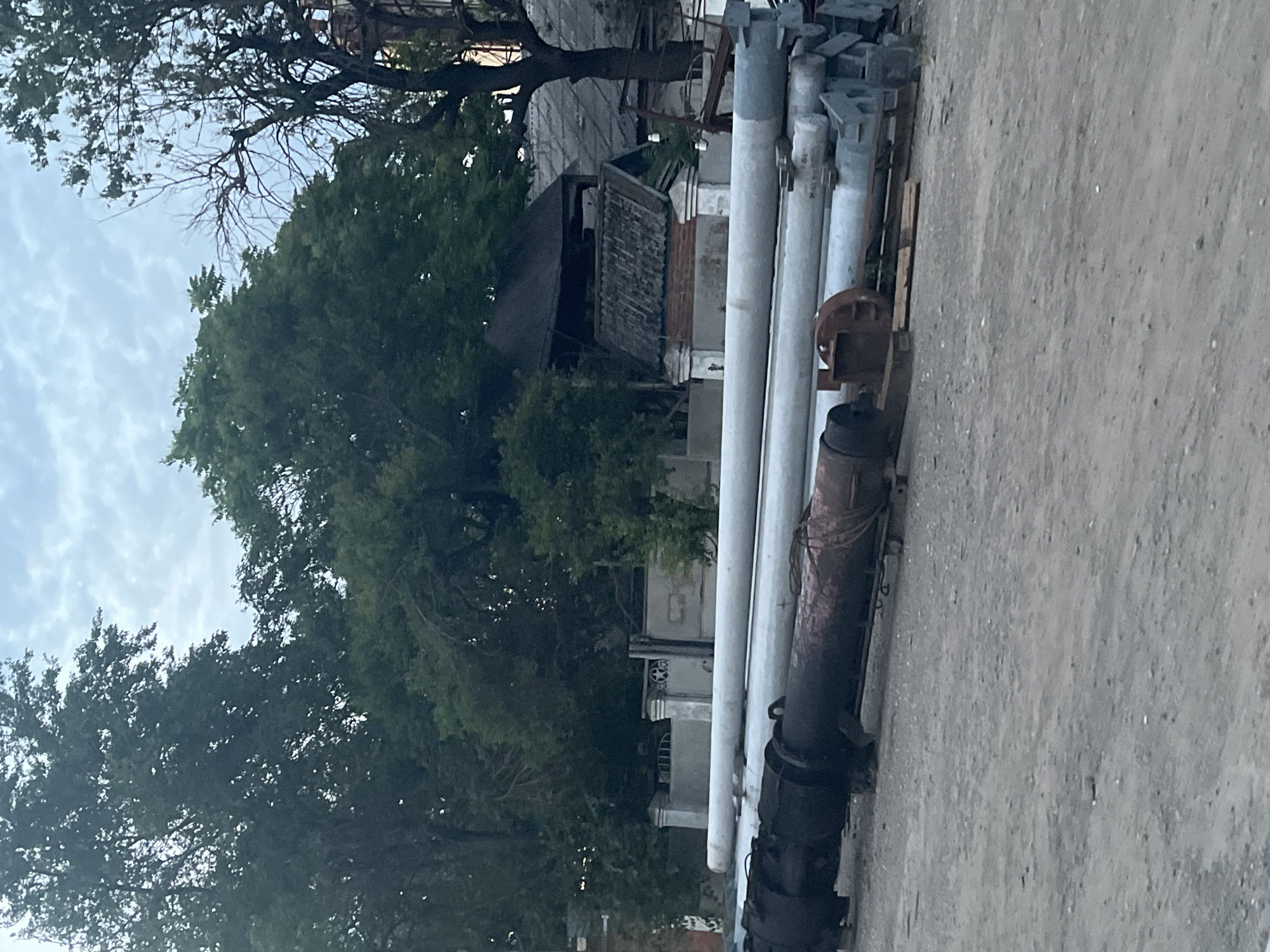 The image shows a row of large, industrial pipes, likely made of concrete or metal, laid out on the ground. Some of the pipes are plain gray, while others are darker and appear older, possibly showing signs of rust or wear. Attached to some of the pipes are metal components, resembling parts of machinery or support structures. The pipes are arranged horizontally in the foreground, filling a significant portion of the frame.
Behind the pipes, a densely leaved tree dominates the mid-ground, its branches reaching upwards and partially obscuring buildings. The buildings in the background appear somewhat dilapidated, with visible brickwork and architectural details suggesting an older structure. The overall scene has an industrial or construction-related feel, possibly a storage yard or a work-in-progress area.
The sky above is overcast with visible clouds, indicating a possibly cloudy day. The lighting is diffused, suggesting it is not direct sunlight, so it could be morning, afternoon, or evening under cloud cover. There are no people visible in the image. No discernible text is present. The setting appears to be an outdoor area, possibly in an urban or semi-urban environment given the presence of buildings and industrial materials. The image is oriented vertically, with the pipes running from the bottom right towards the center left.