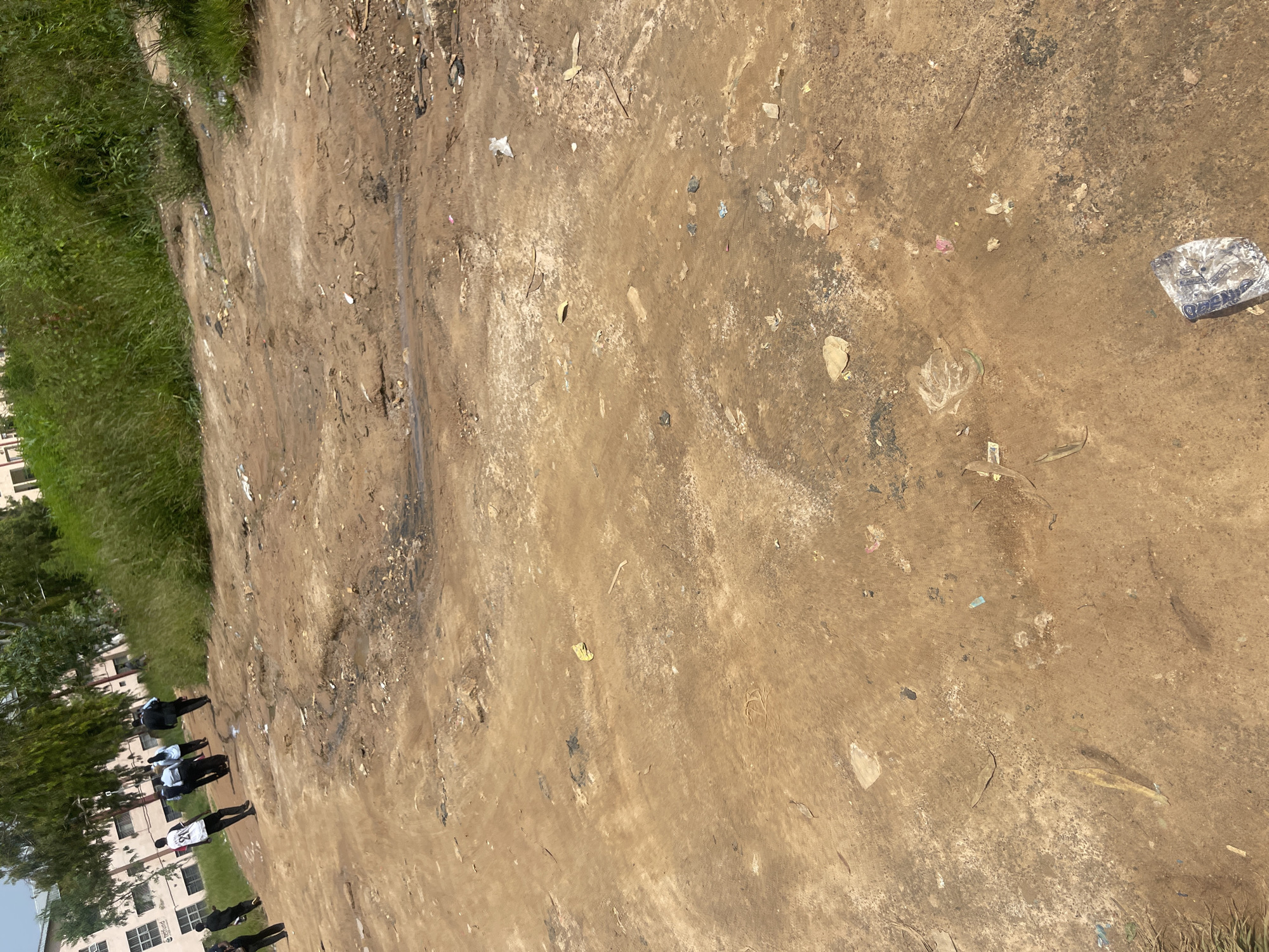 The image shows a group of people walking on a dirt road, with buildings and overgrown vegetation in the background. The ground is uneven and appears to be a construction site or an undeveloped area. There are several scattered pieces of trash on the ground, including plastic bags and wrappers. The people are dressed in casual clothing and appear to be in motion, possibly walking towards or away from the buildings. The overall scene suggests an outdoor environment in a developing area, likely in the city of Jos, Nigeria, given the context.