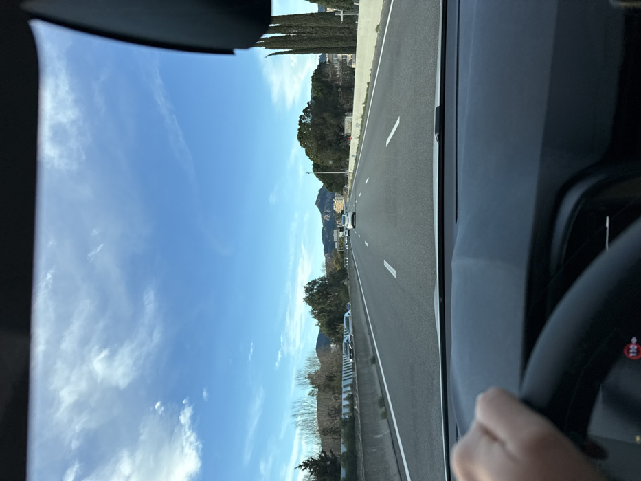 The view from inside a car moving on a highway. The clear blue sky dotted with white clouds is visible through the front windshield. Mountain peaks are seen in the distance beyond the trees lining the road. Cars are ahead on the multi-lane highway. A steering wheel and part of the dashboard are visible on the right side of the frame.