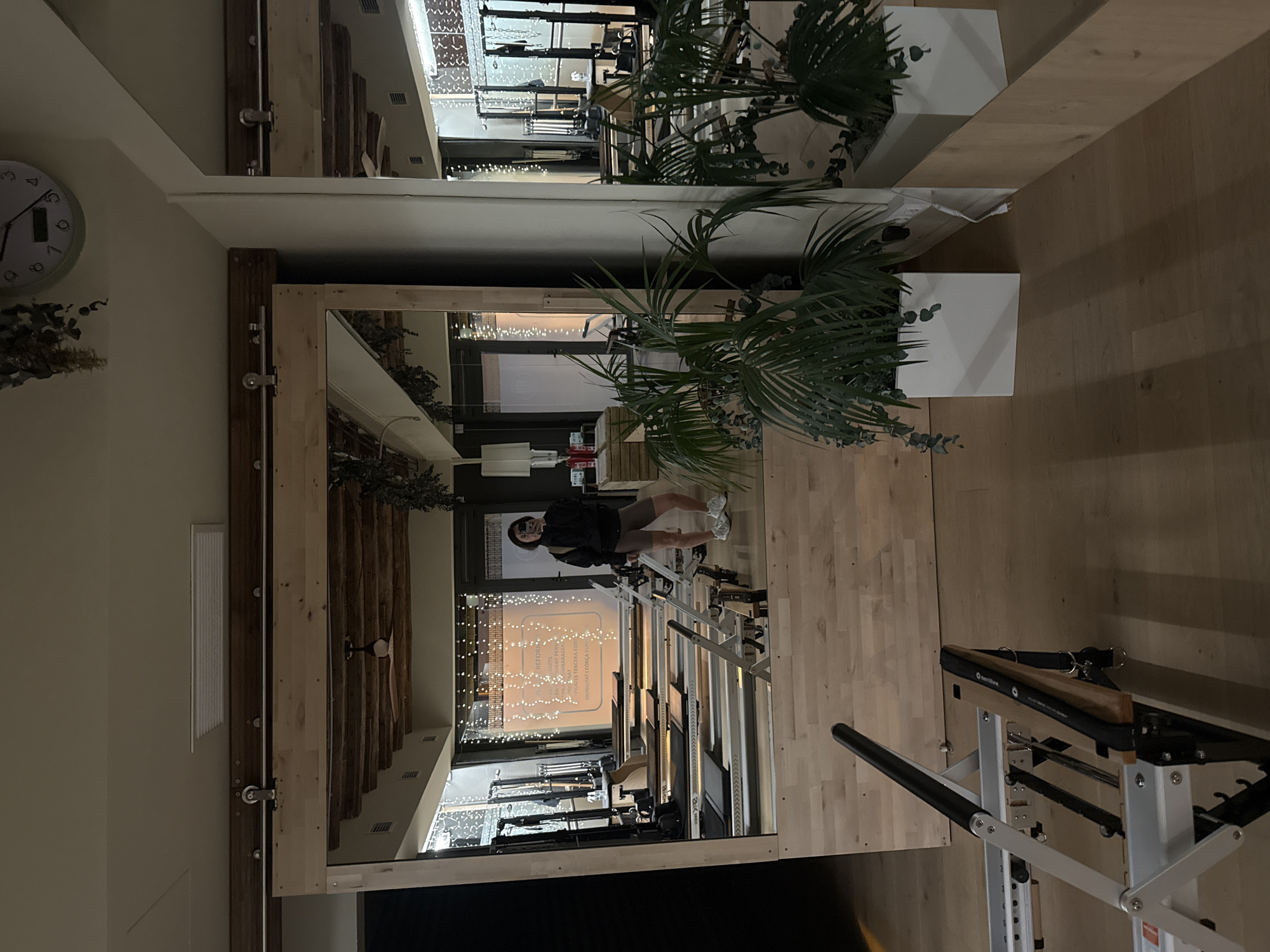 The image shows a reflection in a large mirror within what appears to be a Pilates studio, identifiable by the Pilates equipment. The studio has wooden floors and a wooden frame around the mirror. A person is visible in the mirror, wearing a black jacket and shorts, and standing beside a Pilates reformer machine. The studio's interior is illuminated by string lights hanging in front of a window. Text is visible on the window indicating that the studio offers Pilates services in Barcelona. Plants in white square containers are placed near the mirror. A Pilates Cadillac machine is also visible in the studio. A clock is seen on the wall next to the mirror.