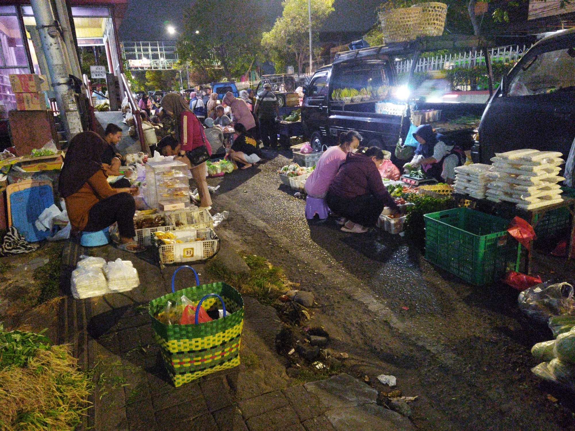This is a nighttime street scene in Semarang, Indonesia, likely a market area. Several people are present, engaged in various activities. In the foreground, a woman wearing a hijab and an orange top is seated on a blue stool, interacting with produce. To her left, a pile of green leafy vegetables is on the ground. A large green and yellow woven basket sits nearby, containing red bags and other items.
Further back, there are several parked vehicles, including a dark van and other vans or trucks, suggesting transport for goods. People are gathered around these vehicles, some appearing to be vendors and others customers. There are stacks of white packaged goods, possibly food items, on display or for sale. Baskets and crates are used to hold and display produce. The ground is wet and appears to be asphalt or a similar paved surface, indicating recent rain or cleaning. The overall lighting is artificial, with streetlights and vehicle headlights illuminating the scene. The environment seems informal, typical of a street market at night.