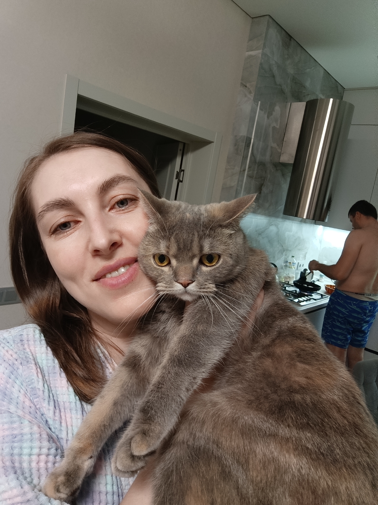 A woman takes a close-up selfie while holding a large, gray and brown cat, set in a kitchen in Chaikovsky, Russia. The woman, with light brown hair and blue eyes, offers a gentle smile to the camera. She is wearing a light-colored, possibly pastel-striped or checkered, long-sleeved top. The cat, with striking yellow eyes and prominent white whiskers, appears to have an unamused or indifferent expression while being held.
In the background, a man, shirtless and wearing blue patterned shorts, is seen from behind, standing at a kitchen stove and cooking in a black pan. Above the stove, a modern stainless steel range hood is visible, and the kitchen backsplash is a light, possibly marble-patterned material. Bottles are also visible on the countertop next to the stove. The scene is brightly lit, likely indoors during the day. No visible text is present in the image.