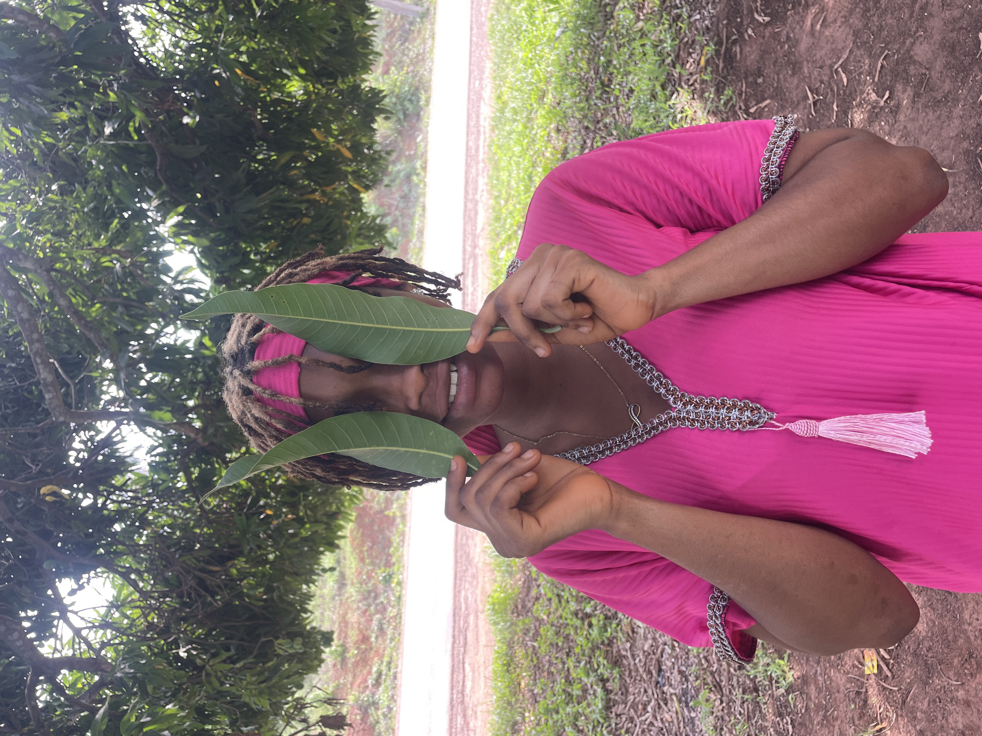 The image displays a person outdoors, captured in a playful moment under a lush tree canopy, likely in Ogbe, Nigeria. The individual, with medium-dark skin and dreadlocks, is wearing a vibrant hot pink tunic or dress. This garment features an elaborately embroidered neckline with silver-toned chain-link details, pink stitching, and a decorative pink tassel hanging from the front. They also sport a pink headscarf.
The central activity involves the person holding two large, green leaves – reminiscent of mango leaves – one over each eye, partially obscuring their face. A clear, happy smile is visible beneath the leaves, conveying a joyful or mischievous mood. Their hands, with well-manicured nails, are visible holding the leaves. Around their neck, a delicate silver-colored necklace with a distinctive pendant can be seen.
The setting is an unpaved, natural outdoor environment. Densely packed green foliage dominates the background and upper sections of the image, suggesting the person is standing under a large tree. A dirt path or road is visible further back, separating areas of green grass and scattered dry leaves on the ground. The lighting indicates it is daytime, with sunlight filtering through the leaves, creating soft highlights and shadows. No visible text is present in the image.