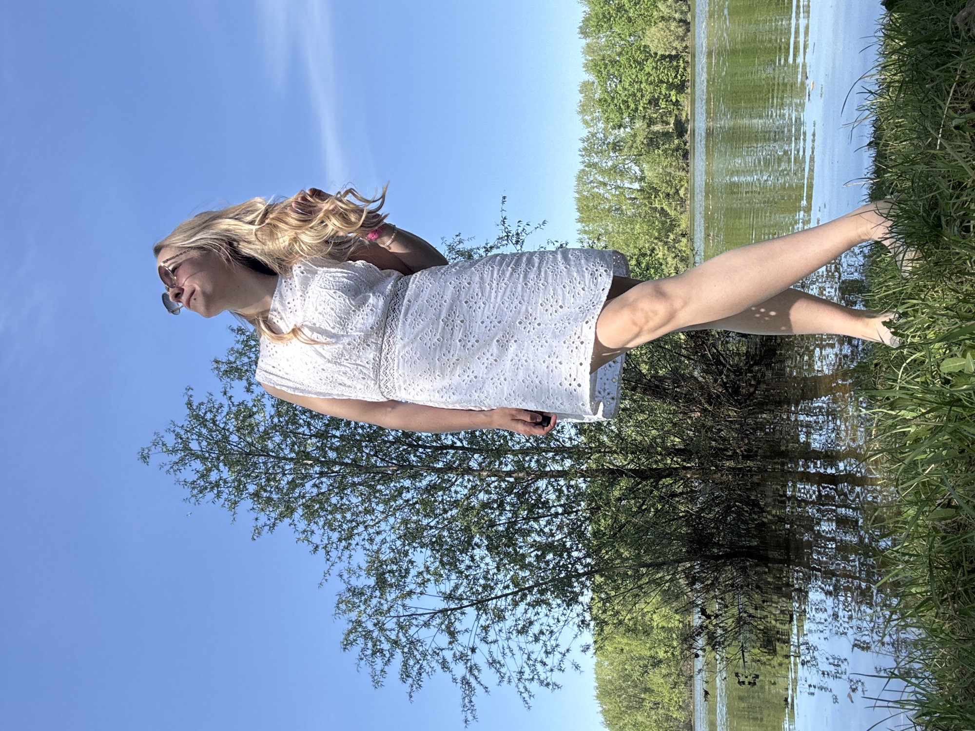 A woman wearing a white dress and sunglasses stands near a lake, likely in the city of Chaikovsky, Russia. She is partially immersed in the water, with her bare feet visible. The background includes trees and a blue sky. She is holding a black object in her hand.
