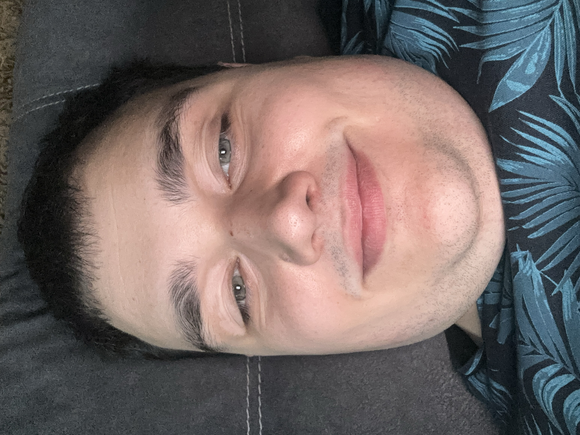 This is a close-up, overhead, vertical shot of a person's face. The person appears to be lying down, with their head angled slightly upwards towards the camera. Their skin is pale, with some redness on the cheeks and a few stray facial hairs visible on the chin and jawline. They have dark, slightly messy hair that frames their face.
The person's eyes are open and looking towards the camera, with one eye more prominently in view. The eyes are a light grey-blue color. The eyebrows are dark and well-defined. Their lips are slightly parted and appear to be a natural pink color. There is a subtle smile on their face, suggesting a relaxed or content emotion.
The person is wearing a dark blue shirt with a tropical leaf pattern in a lighter blue and teal. The background consists of a dark grey surface, possibly a couch or bed, with some visible stitching that runs horizontally across the image. The lighting is soft and even, with no strong shadows, suggesting an indoor setting, possibly during the day. There is no visible text in the image.