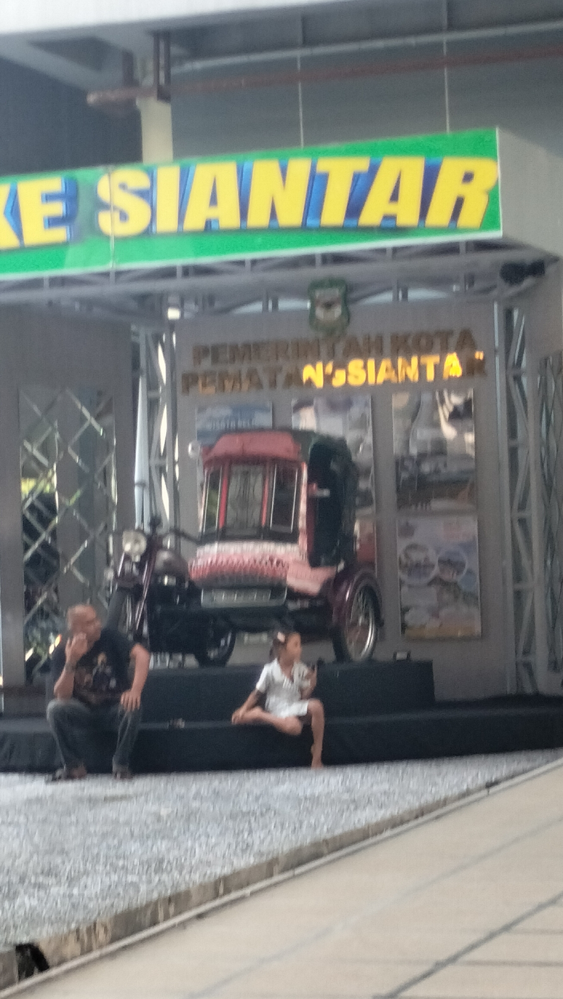 The image captures a street scene in Ramunia Dua, Indonesia, under a bright, possibly overcast sky, with overhead structures suggesting an urban or semi-urban setting. A prominent green banner with yellow lettering, reading "KE SIANTAR", is visible at the top, indicating a directional sign or advertising. Below this, a display features a vintage three-wheeled vehicle, possibly a type of auto rickshaw or modified motorcycle, painted in a dark color with red accents and a fabric canopy. The vehicle is positioned on a raised black platform.
Two individuals are present. To the left, a man is seated in a crouched position, looking to his left with his hand near his face. He is wearing dark clothing. To the right of the vehicle, a young girl is seated on the same black platform, facing towards the left. She is wearing a white outfit and appears to be holding something in her hands. Behind the vehicle, a sign with text that appears to be in Indonesian, including "PEMERINTAH KOTA" and "PEMATANGSIANTAR", suggests the display is related to the local government of Pematangsiantar. Several framed posters or images are mounted on the wall behind the vehicle. The foreground consists of a paved area with visible gravel and a concrete edge.