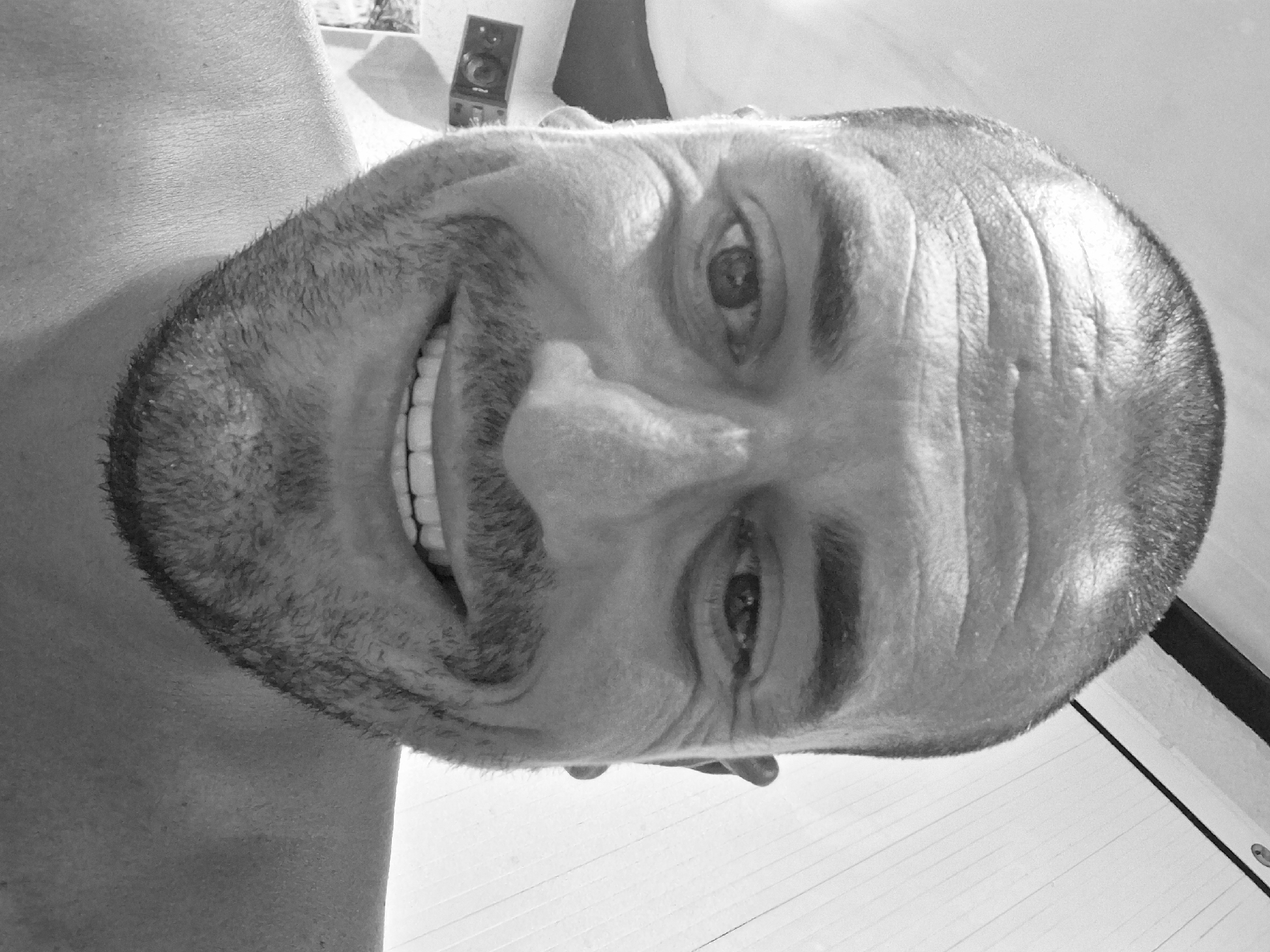 A black and white, close-up portrait depicts an adult male, reportedly in Shkorpilovtsi, Bulgaria. The subject has a closely cropped haircut, pronounced horizontal wrinkles on his forehead, and dark eyebrows. His brown eyes are visible, with the left eye (viewer's right) more prominently featured. A mustache and short, dense beard (stubble) frame his open-mouthed smile, which reveals his upper teeth. His bare shoulders are visible at the bottom of the frame. The background suggests an indoor setting; the upper right reveals a light-colored wall with vertical paneling, a dark horizontal beam, and a small, dark, circular wall fixture. The upper left background includes a darker wall area with a wall-mounted rectangular object containing a circular element, possibly a speaker or camera, and a lighter rectangular object positioned below it.