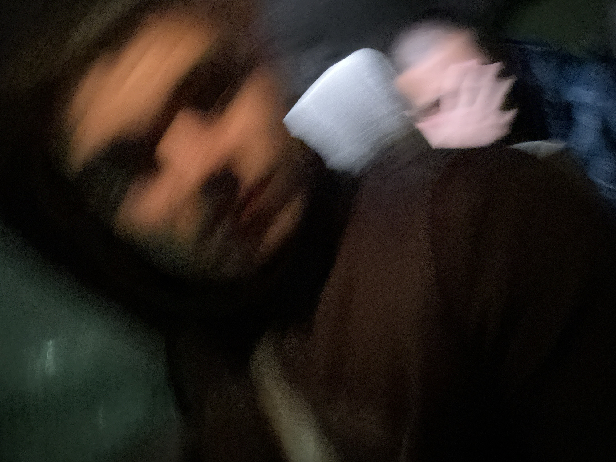 The image displays a heavily motion-blurred and underexposed scene, located in the city of Unknown, Unknown. A single individual is visible, centered in the frame, their features rendered indistinct due to blur and low light. The subject's face appears in the upper right portion, showing an outline of a nose and mouth, suggesting a medium skin tone and dark hair or head covering. The subject wears a dark, possibly brown, garment covering the torso. Below the torso, two light-skinned hands are visible, appearing clasped or positioned closely together, also significantly blurred. A small, irregularly shaped light-colored (white or light grey) object is present near the subject's midsection, between the torso and the hands. The lower-left area shows blurred blue fabric, consistent with denim trousers. The background is dark and indistinct, with a faint greenish-teal area in the upper left quadrant containing horizontal streaks of lighter tone. No specific architectural elements or environmental details are discernible to precisely identify the setting within the city.