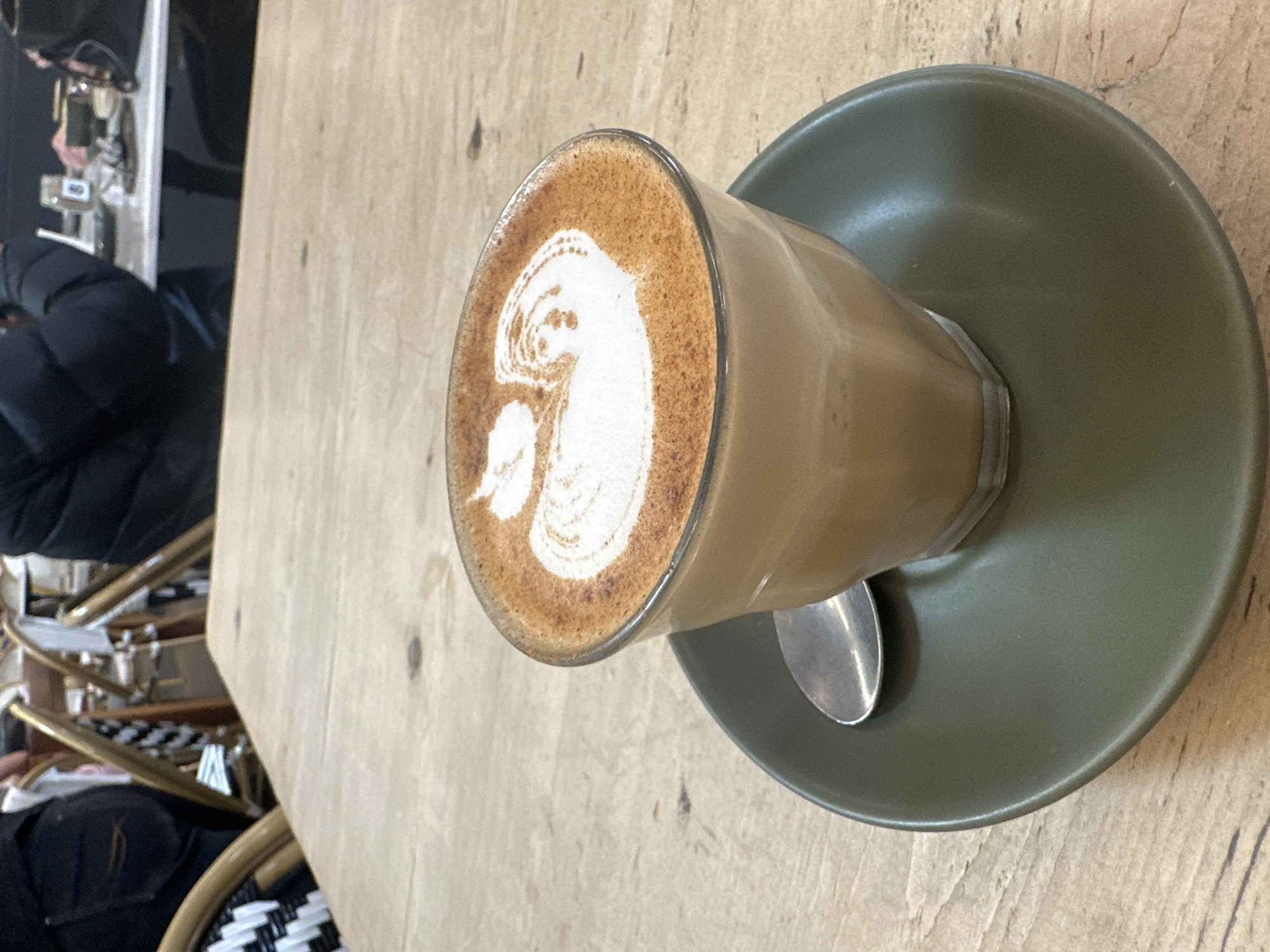 A close-up shot features a glass of coffee with latte art, resting on a dark green saucer. The coffee has a foamy top with a white pattern. A silver spoon is placed on the saucer. The coffee cup is on a light-colored wooden table. In the background, people are seated at tables and chairs. One person is wearing a black puffer jacket, and another is wearing black pants. Chairs with woven seats and backs are visible. The overall scene suggests an indoor cafe setting.