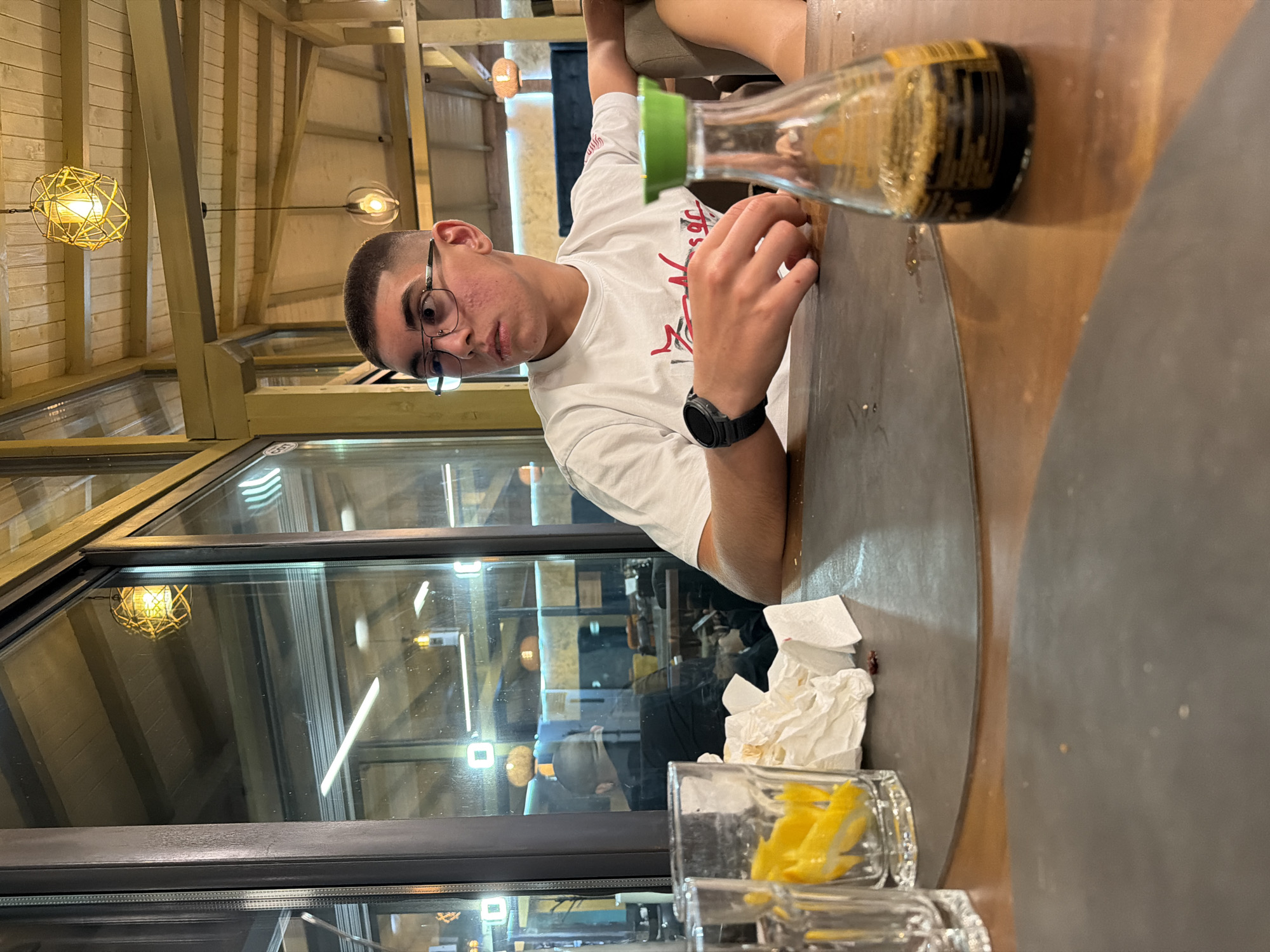In Panagyurishte, Bulgaria, a young male with short hair and glasses is seated at a table in what appears to be a restaurant with a wooden ceiling and yellow accents. He is wearing a white t-shirt and a black watch on his left wrist. In front of him, on the table, are two clear glasses, one containing lemon slices, a crumpled white napkin, and a small glass bottle with a green cap, filled with a dark liquid, likely soy sauce. The background reveals large glass windows reflecting the interior lighting and a glimpse of another person. The lighting in the establishment is warm, with decorative pendant lamps visible.
