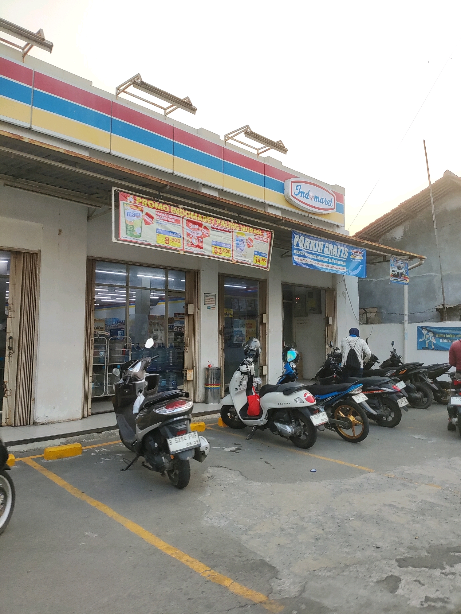 The image shows the exterior of an Indomaret convenience store in Wanajaya, Indonesia. The store has a white facade with red, blue, and yellow stripes across its upper section, and the Indomaret logo is prominently displayed. In front of the store, a row of parked motorcycles is visible on asphalt. Yellow parking lines are painted on the ground. Above the entrance, promotional banners are hung, including one advertising "PROMO INDOMARET PALING MURAH" and another with "PARKIR GRATIS" (free parking). The sky is overcast. A person wearing a white hoodie and backpack is standing near one of the motorcycles.