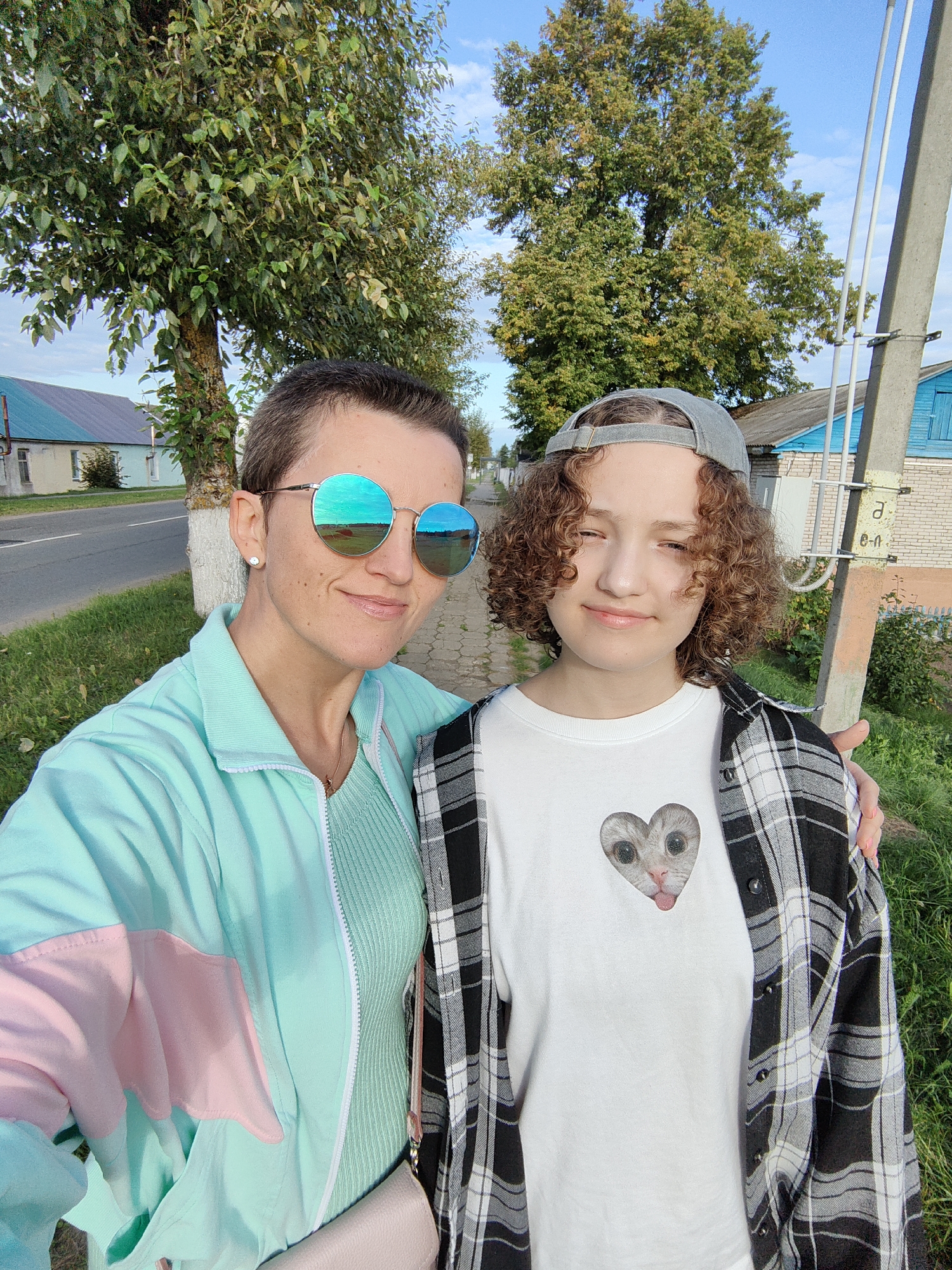 Two individuals, an adult and a younger person, are posing for a selfie outdoors in Myadzyel, Belarus. The adult, on the left, has short brown hair, is wearing large round mirrored sunglasses, a light blue and pink windbreaker, and a mint green ribbed top. The younger person, on the right, has curly brown hair, is wearing a grey baseball cap, a white t-shirt with a heart-shaped graphic of a cat's face, and a black and white plaid shirt worn open. They are standing on a sidewalk next to a grassy area with trees and a road in the background. A light-colored utility pole is visible to the right of the younger person. The sky is blue with some clouds. Two houses, one light yellow with a teal roof and another brick house with a blue roof, are visible in the distance behind them.