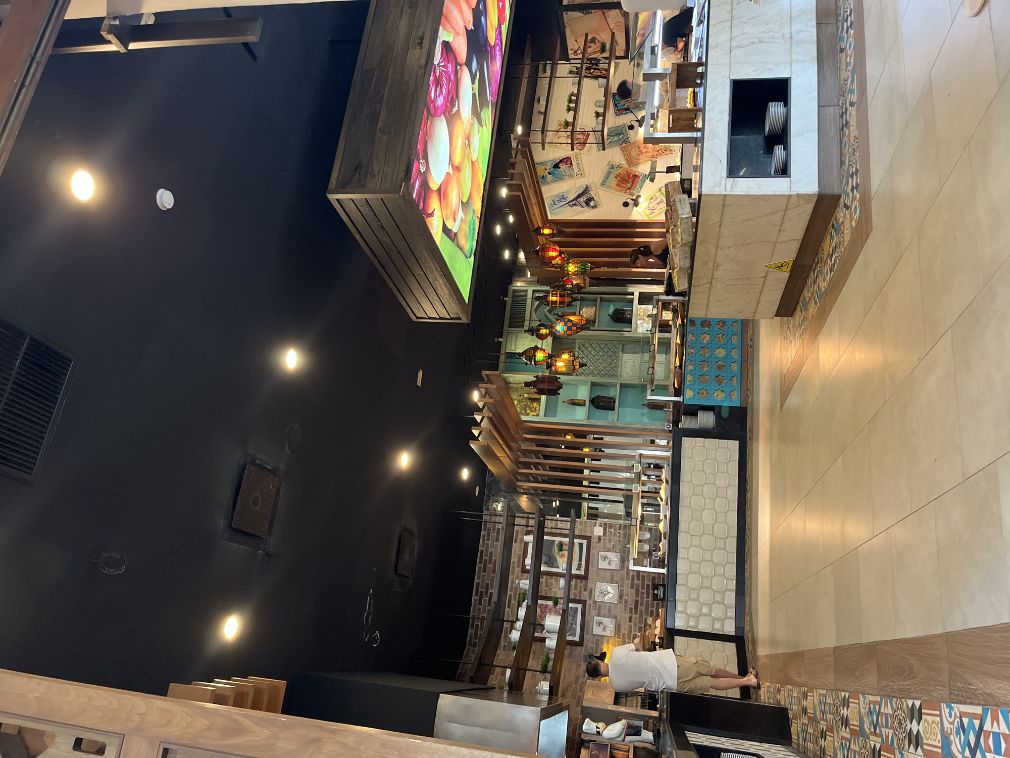 The image depicts an interior view of a restaurant in Playa del Carmen, Mexico. The ceiling is dark, and lighting fixtures provide illumination. A large rectangular display showcases food imagery. The restaurant features diverse decorations, including lanterns, artwork, and brick accents. Tiled flooring transitions into a patterned border. A person wearing a white shirt and khaki shorts is partially visible at the edge of the frame. A counter with visible plates and tiled patterns stands prominently in the foreground.