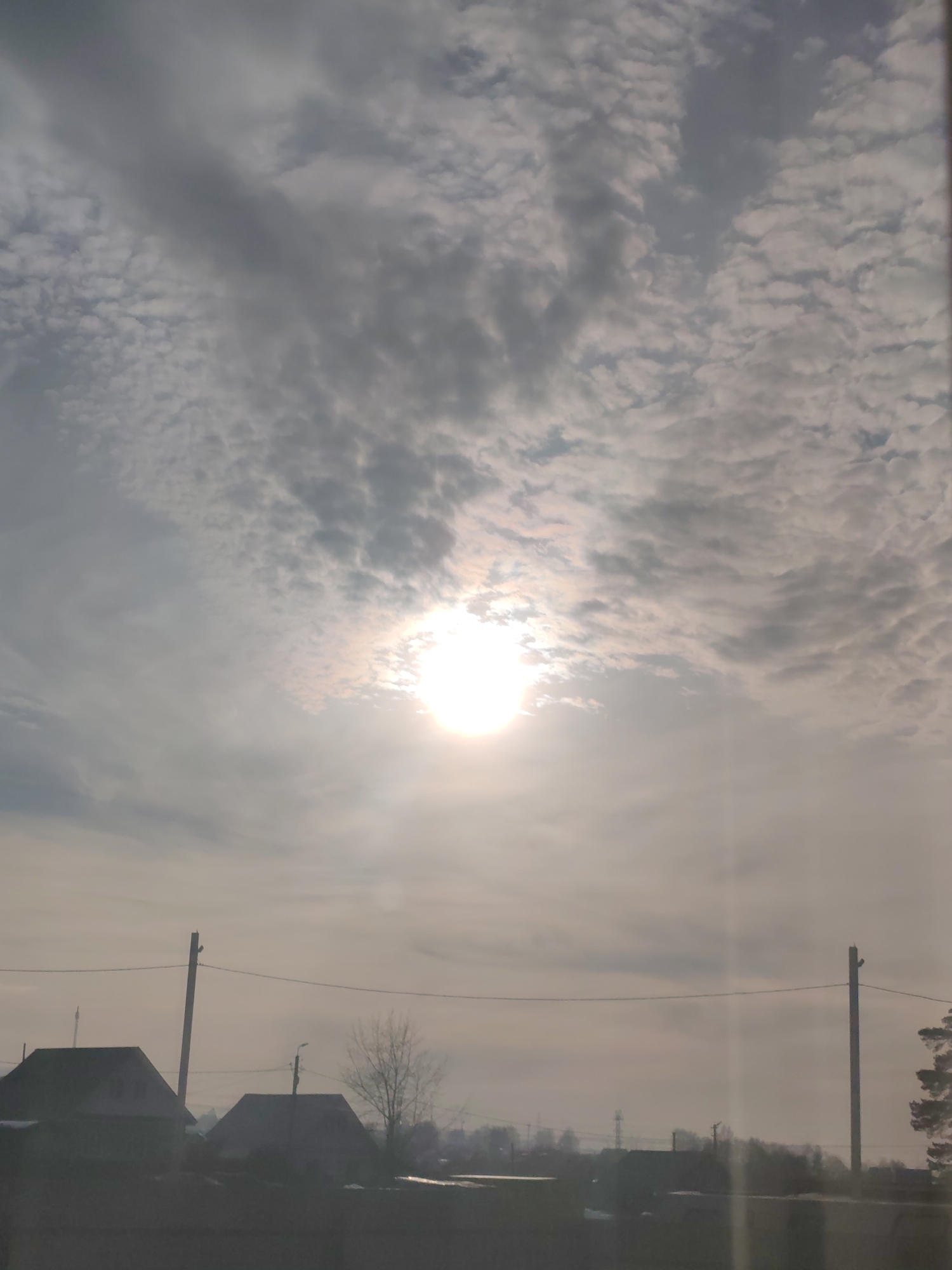 This image depicts a hazy, semi-rural landscape in Chaikovsky, Russia, observed during daylight hours. The upper two-thirds of the frame are dominated by a sky filled with a mix of patchy altocumulus and stratocumulus clouds, presenting a mottled grey-white appearance. The sun is visible as a bright, overexposed circular disc, partially obscured by clouds, positioned slightly above the horizontal center and left of the vertical center. Its light generates a prominent vertical lens flare extending downwards. The lower third of the image shows a blurred horizon with the dark silhouettes of several detached houses featuring sloped roofs, indistinct due to distance and atmospheric haze. Two utility poles, dark against the lighter background, are positioned in the mid-ground; one approximately one-third from the left edge and the other near the right edge, with overhead wires spanning horizontally between them. A bare deciduous tree stands between the poles, and further distant, sparse vegetation is visible. A dark, indeterminate line extends across the bottom foreground, possibly indicating a fence or ground elevation. No people or discernible actions are present.