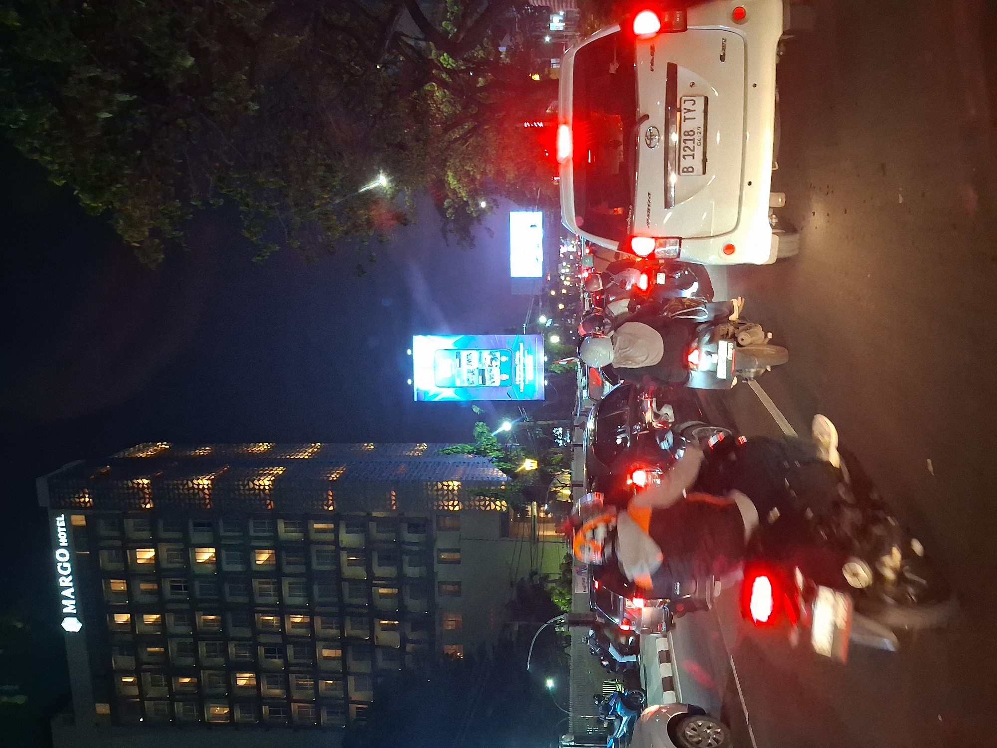 The image depicts a nighttime street scene in Depok, Indonesia, characterized by heavy traffic congestion. A tall, modern hotel, identified as "MARGO HOTEL" by its illuminated signage, dominates the background on the left. The hotel's facade features numerous windows, many of which are lit, giving the building a warm glow against the dark sky.
The foreground and midground are filled with vehicles, predominantly motorcycles and cars, indicating a traffic jam. Several motorcyclists, wearing helmets and riding gear, are visible, some with their headlights and taillights illuminated. One motorcyclist in the foreground is wearing a distinctive orange and white jacket. Cars are also present, with their brake lights glowing red, a common sight in heavy traffic. One white car, a Toyota Avanza, is prominently featured, displaying a license plate that reads "B 1218 TYJ" and a registration sticker indicating "04-29".
Large digital billboards are visible in the midground and background, one displaying bright, colorful imagery. Trees line the right side of the street, their dark foliage contrasting with the artificial lights of the city. The overall atmosphere suggests a busy urban environment at night, with the usual hustle and bustle of traffic. The presence of multiple illuminated signs and buildings points to a commercial or downtown area.