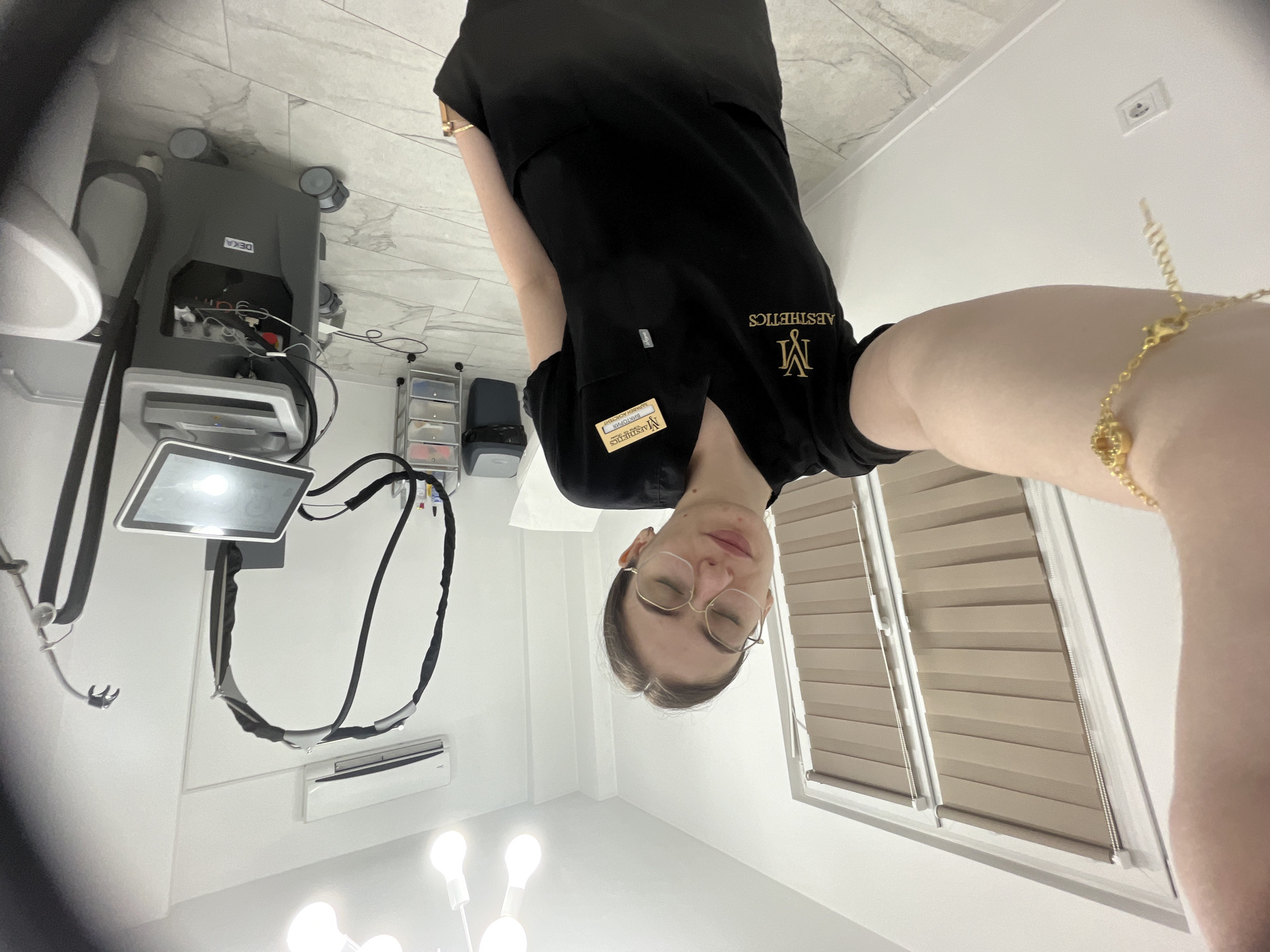 An image, rotated 90 degrees clockwise, shows a person (likely female) from a low-angle perspective, taking a selfie. The person is wearing a black short-sleeved scrub top, which features gold embroidery of a stylized "M" and the word "AESTHETICS". A name tag on the left pocket reads "MAESTHETICS", "BARTOLOVA", and "LASER ACADEMY". She wears glasses and a gold bracelet on her right wrist. Her eyes are partially closed, suggesting a relaxed pose.
The setting appears to be a professional medical or aesthetic clinic. In the background, a large, grey piece of equipment with wheels is visible, bearing the brand name "DEKA". This machine has several cables and hoses connected to it. Adjacent to the equipment is a monitor with a bright screen. A small, clear plastic drawer cart is also present.
The room has white walls, a light-colored tiled floor, and a window with light brown horizontal blinds. A wall-mounted air conditioning unit is installed. Bright overhead lighting from a multi-bulb fixture is visible at the bottom of the frame. The overall impression is a clean, well-lit treatment room.