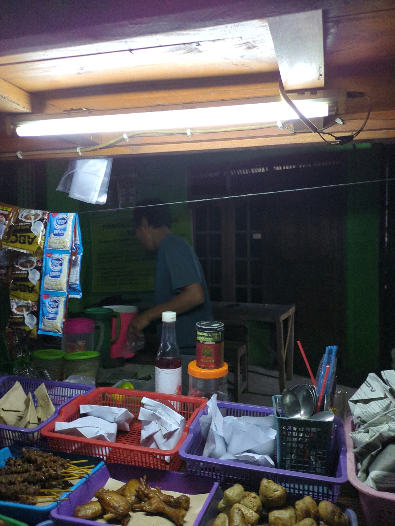 A person, likely a vendor, stands behind a food stall in Sindon, Indonesia. The stall is illuminated by fluorescent lights. In front of the vendor, there are several baskets and containers holding food items. One basket contains skewers of cooked meat, another has what appears to be fried chicken pieces and other small fried items. There are also packets of powdered drinks hanging on a rack, and various containers with lids, including a green and pink pitcher, and a glass bottle with a white cap. In the foreground, a red basket holds white paper-wrapped parcels. A dark wooden table and chair are visible in the background, as well as a green wall.