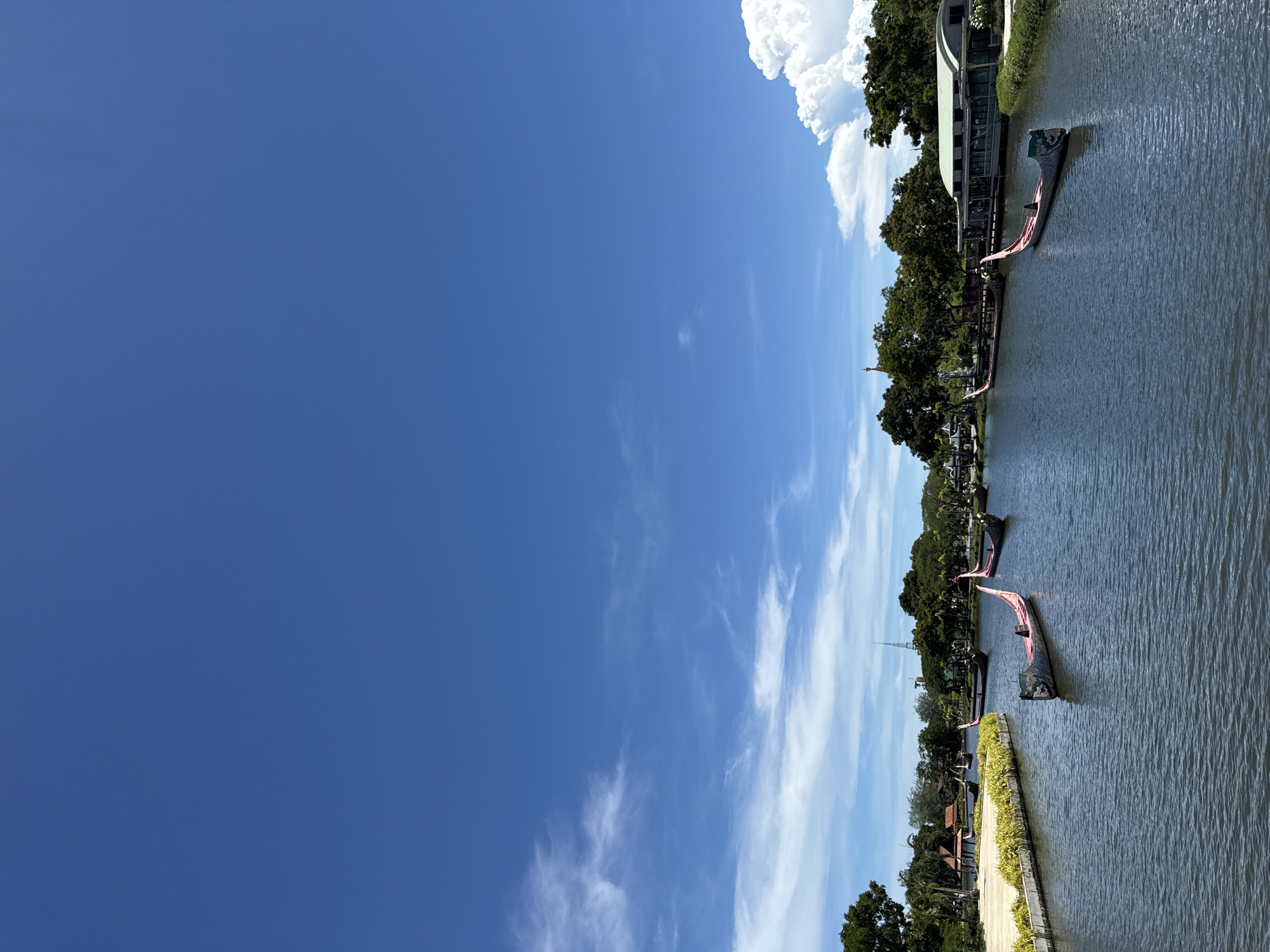 The image depicts a waterway in Bang Pu Subdistrict Municipality, Thailand, under a bright blue sky with scattered clouds. The water surface is rippled and calm. Along the waterway, there are trees, vegetation, and architecture resembling buildings. Gondola-like boats, decorated with red accents, are docked along the edge.