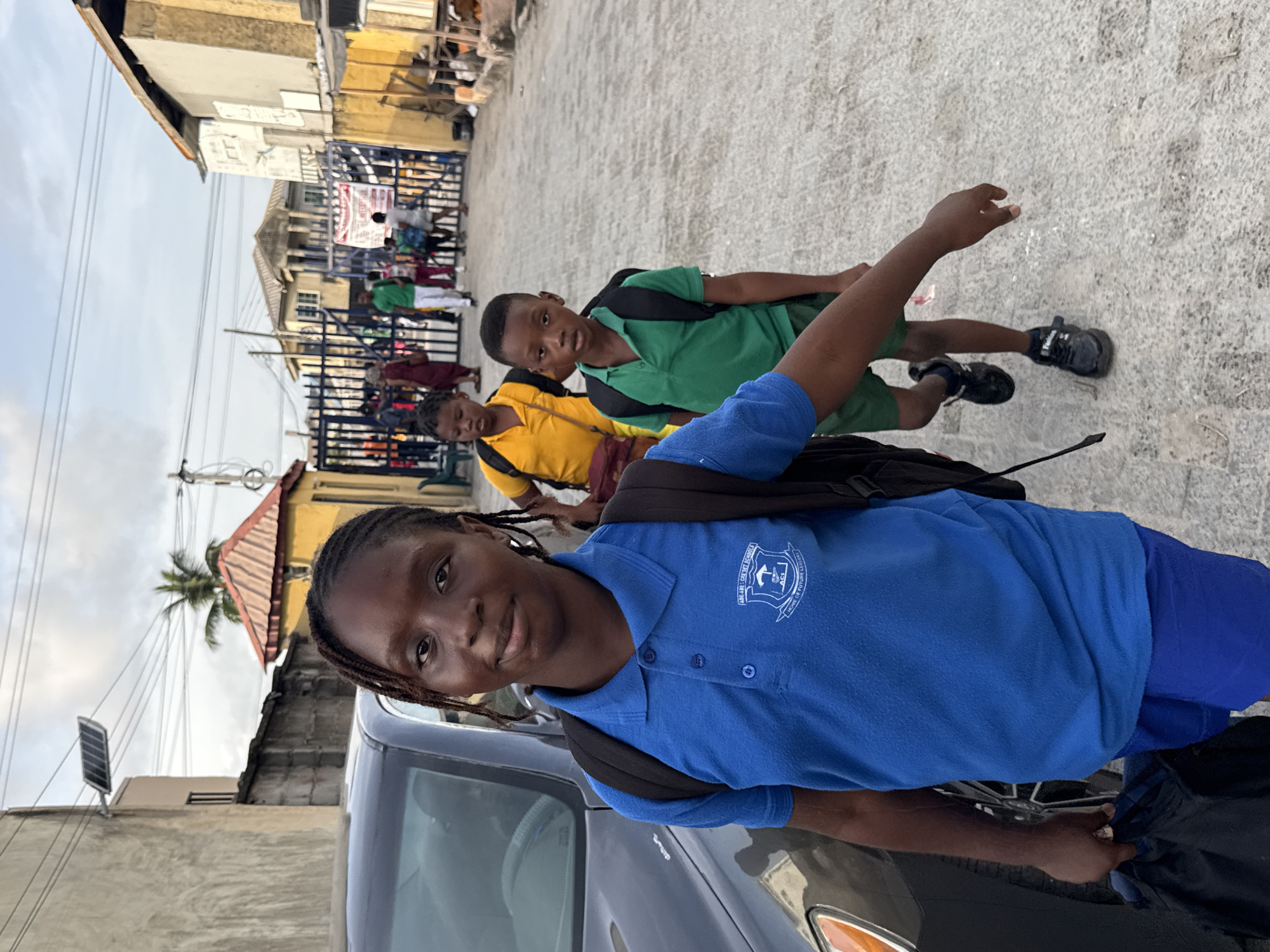 A young girl in a blue school uniform stands near a gray car on a textured gray concrete street in Ado, Nigeria. Behind her are two younger children in yellow and green uniforms, also with backpacks. In the background, a blue metal fence surrounds a crowded area with more people in school uniforms. Buildings of varying heights and colors line the street. A palm tree is visible among the buildings. The sky is overcast.
