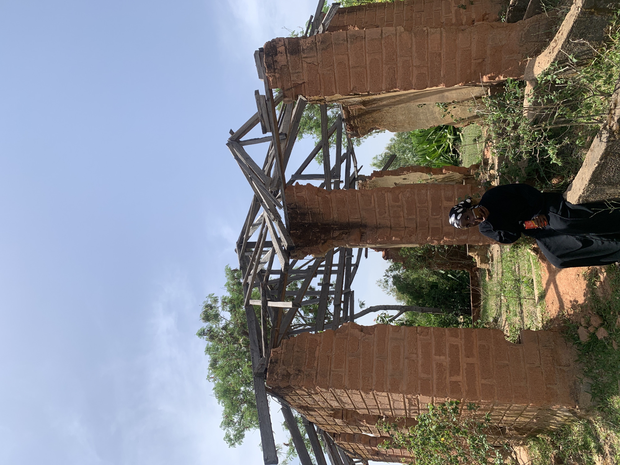 The image depicts the crumbling remains of a brick structure, likely a building, against a backdrop of a pale blue sky and some greenery. The structure appears to be in a state of disrepair, with sections of the brick walls missing and a decaying wooden framework overhead. A person is standing to the right of the frame; wearing a black garment with a patterned headscarf. The person is holding an object near their ear, possibly a mobile phone. The environment surrounding the building is green, suggesting vegetation or foliage. The location is Gwafan, Nigeria.