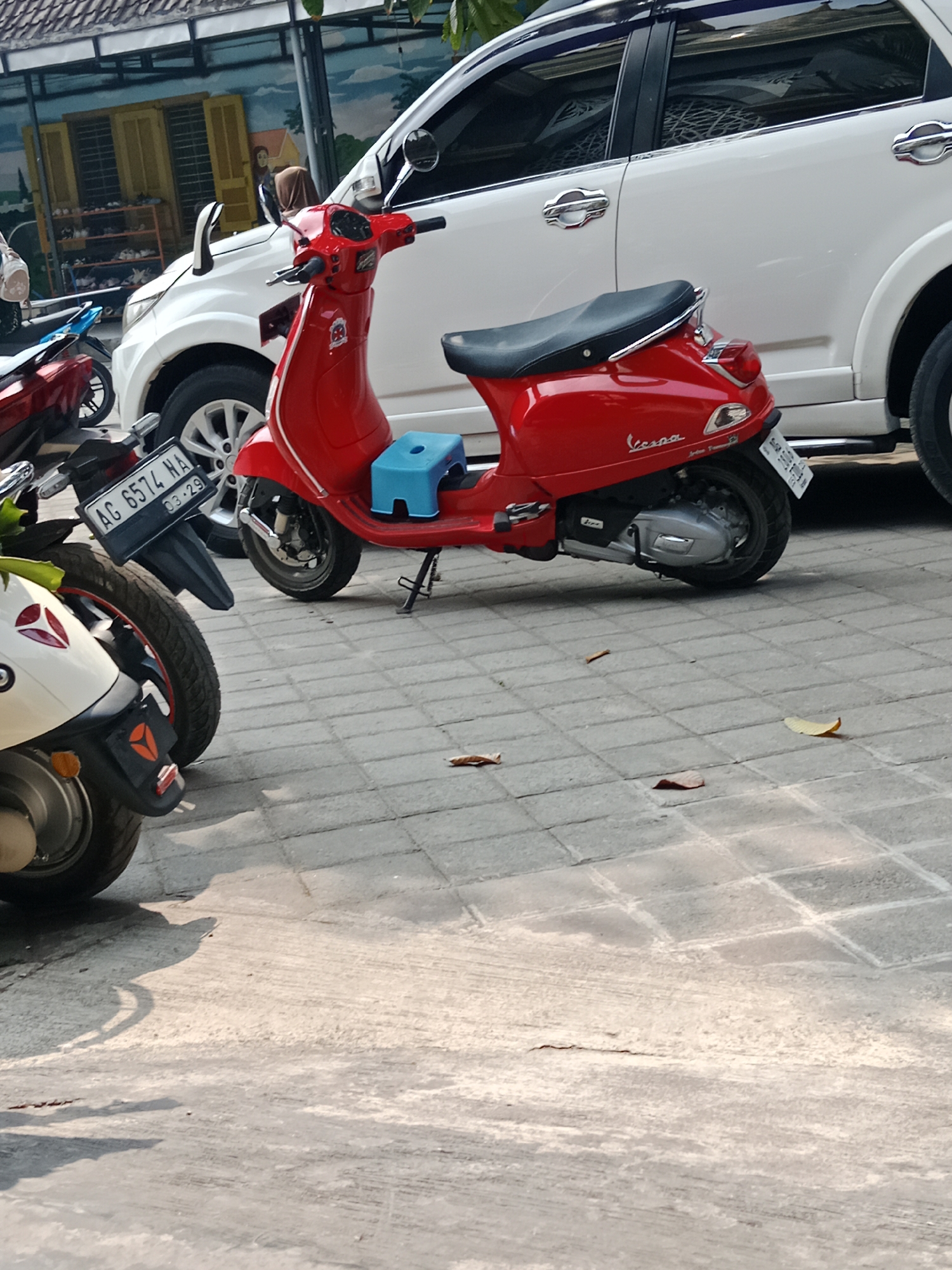 Two scooters are parked on a paved surface in front of a white vehicle, likely a car. The scooter in the foreground on the left is partially visible and appears to be white and red. The scooter in the middle is red and has a blue plastic stool on its seat. Its license plate reads "AG 6574 HA 03-29". The white vehicle is parked behind the scooters. In the background, there is a building with yellow doors and a mural on the wall. The environment suggests an outdoor setting in a city, possibly a parking area or street in Srikaton, Indonesia, given the license plate format. The lighting indicates it is daytime.