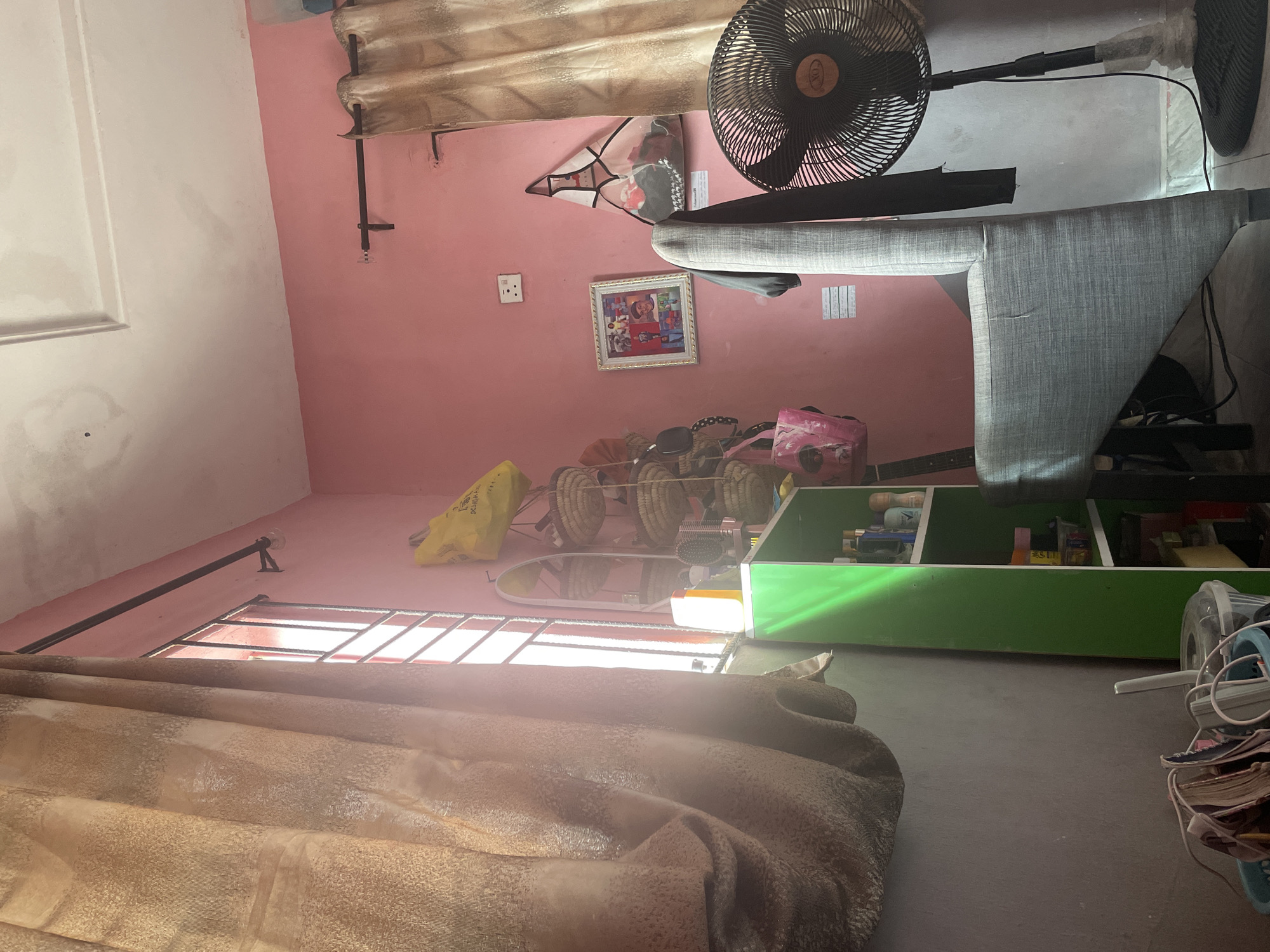 This image depicts the interior of a room in Iyafin, Nigeria. The room is painted pink and features a window with bars covered by a patterned beige curtain on the left. A green shelf unit filled with various items, including bottles and containers, stands against the wall. On the right side of the room, another beige curtain hangs, and a black standing fan is positioned near the wall. A grey upholstered chair is placed in the foreground, partially obscuring the fan and the shelf. On the pink wall, a framed picture hangs, along with a white electrical outlet. Several woven baskets are suspended from the wall, and a yellow bag is placed on top of them. A clear plastic bag containing items is also visible. The floor appears to be tiled.