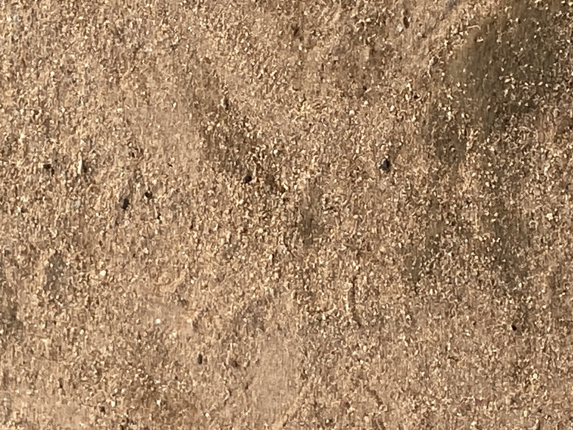 The image is a close-up, high-angle shot of dry, sandy ground. The sand is a light brown color and appears to be composed of small grains and pebbles. There are subtle variations in texture and color across the surface, suggesting it might be a path, a playground, or an open area in Jauro Yinu, Nigeria. No people, subjects, objects, or actions are clearly discernible in the image; it is solely focused on the ground's texture and composition. The lighting suggests an outdoor setting, likely under natural sunlight, casting soft shadows that highlight the granular nature of the terrain.