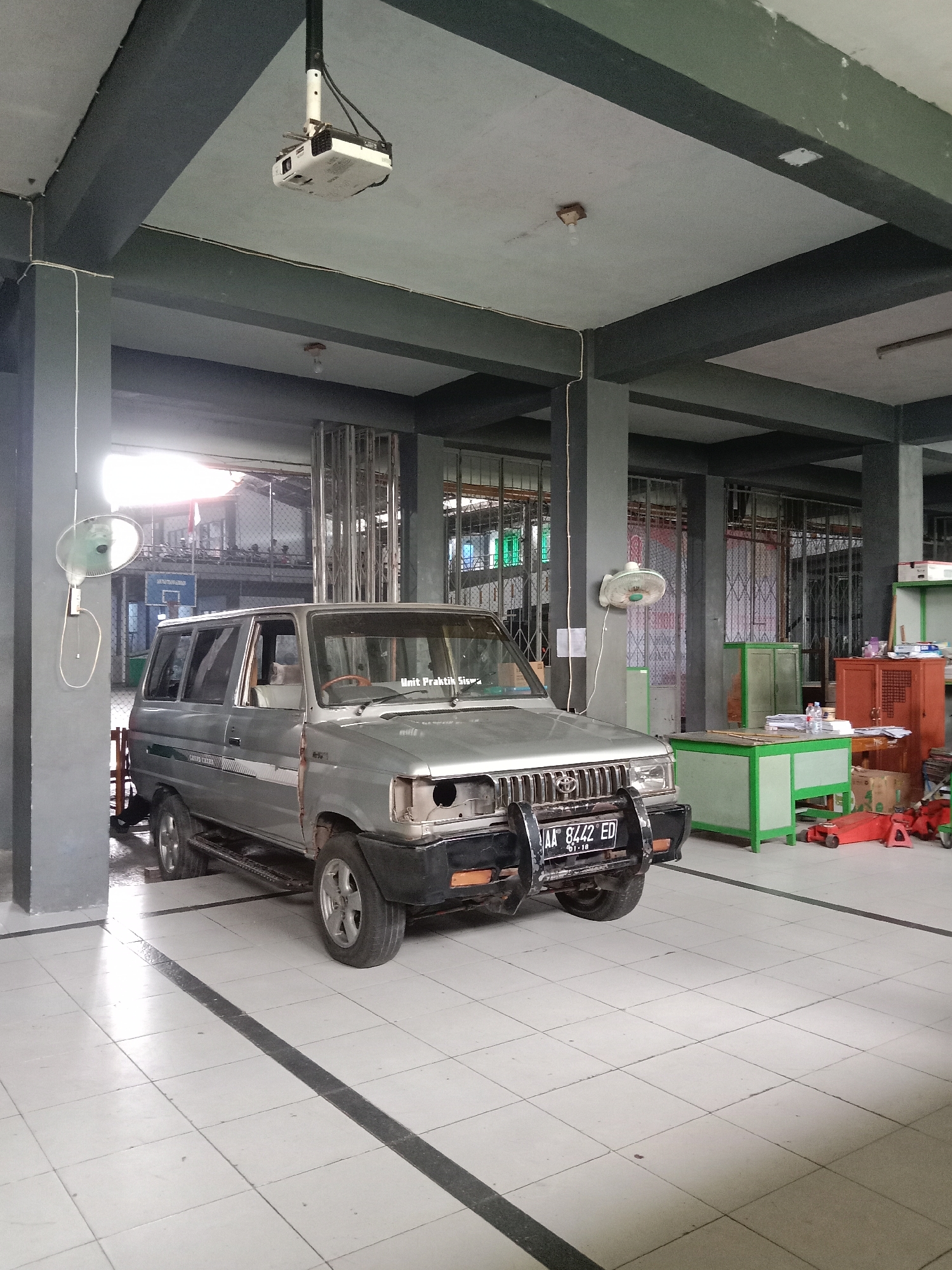 The image depicts an indoor scene, likely a garage or workshop, with a silver van parked in the foreground. The van has a license plate that reads "AA 8442 ED 01-18". The interior features gray concrete pillars and a white tiled floor with dark gray grout lines. On the ceiling, a white projector is suspended. To the left of the van, a white oscillating fan is mounted on a gray pillar. In the background, there are green cabinets, an orange cabinet, a green desk with papers and a bottle of water, and some red car jacks. The overall impression is of a functional space, possibly associated with a vocational school or repair facility, given the presence of a projector and what appears to be automotive equipment. The mention of Kebumen, Indonesia, provides a geographical context for this scene.