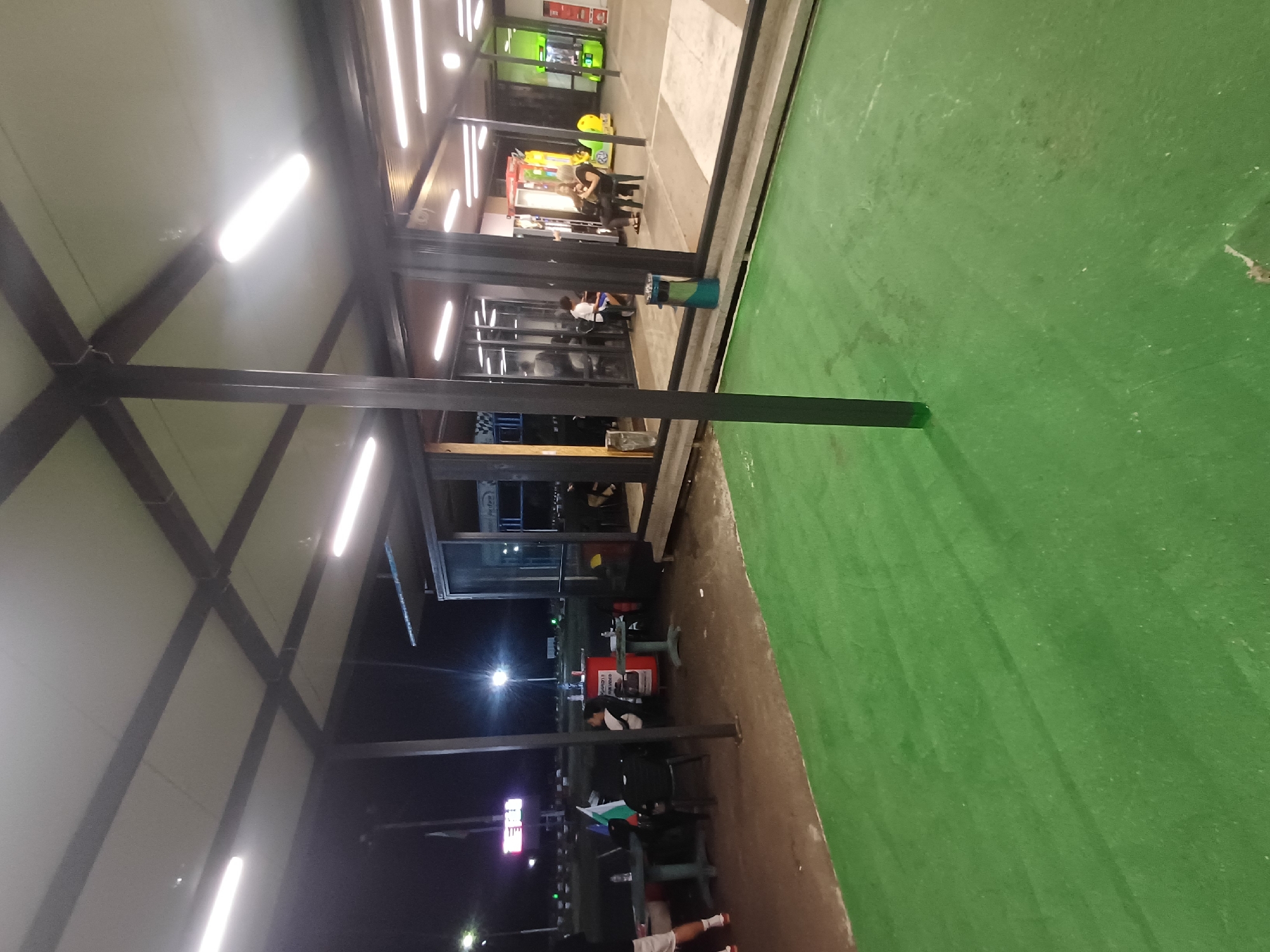The image depicts an outdoor seating area with a green artificial turf surface in front of a modern building with large glass windows and an overhang. The scene appears to be at night, with artificial lighting from fluorescent tubes illuminating the covered area and the interior of the building. Several people are visible seated at tables outside, some with Bulgarian flags displayed. Inside the building, more people are seen, and arcade-like machines are visible, suggesting a recreational or entertainment venue. The background shows a dark sky with some distant lights and what appears to be a score display board. The location is identified as a karting track in Varna, Bulgaria.
