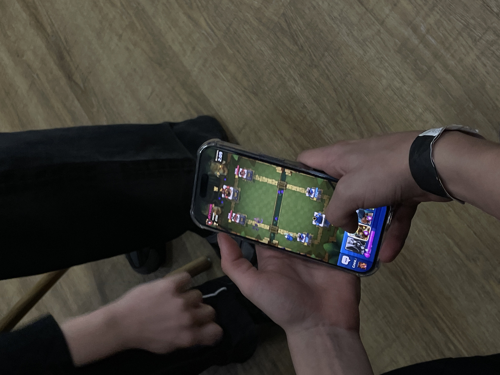 An indoor scene in Krasnoyarsk, Russia, depicts a smartphone actively displaying the mobile game "Clash Royale." The phone, identified as an iPhone within a clear protective case, is held vertically by two hands of an individual. The game screen shows a battle in progress with multiple virtual units deployed on the field and a timer in the upper right corner indicating "2:09." The lower portion of the screen displays playable game cards and an elixir resource bar. A black and white event-style wristband is present on the left wrist of the person holding the phone. In the upper left background, the forearm and hand of a second individual are visible, wearing dark long-sleeved clothing, with fingers extended towards the area of the phone. A segment of a light-colored wooden object, consistent with a furniture leg, is partially visible behind the second individual's hand. Dark-colored trousers and footwear belonging to individuals are discernible in the background. The floor consists of light brown, wood-patterned laminate or planking.