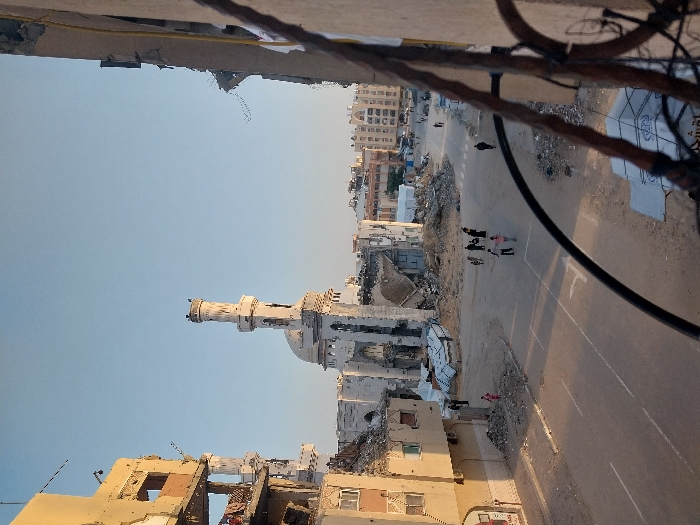 This image captures a heavily damaged urban intersection in Khan Yunis, Palestinian Territory, under a clear, light blue sky. At the center, the Great Mosque of Khan Yunis stands significantly damaged, with its white minaret largely intact but its main structure extensively collapsed and parts of its dome absent. Surrounding the mosque are substantial piles of concrete rubble, twisted rebar, and debris. To the left, a multi-story building, yellowish in hue, shows severe structural compromise, with large sections of its facade missing, exposing internal rooms, and numerous impact holes visible. Another severely damaged building partially frames the right side of the image, viewed through dark, rusted metalwork. A paved road with visible white lane markings, including solid and dashed lines, traverses the foreground and midground. Approximately 10-15 individuals are observed walking or standing on the road and in unpaved areas amid the debris, moving through the scene. Their actions are primarily consistent with pedestrian transit. The ground around the structures is covered in scattered debris and dirt.