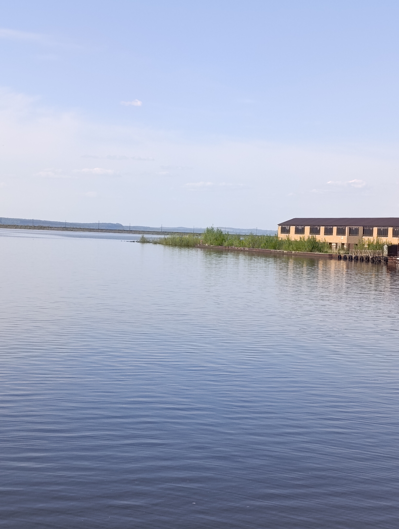 The image shows a body of water, likely a reservoir or wide river, with a large, low building on the right. The building has multiple windows and appears somewhat dilapidated. There are some trees and vegetation growing along the edge of the water near the building. In the distance across the water, there is a line of trees and what appear to be power lines or utility poles. The sky is light blue with some clouds. Based on the presence of the Volga-Kama Cascade of Hydroelectric Power Stations, and the look of the buildings and other characteristics, it is consistent that this may be in the city of Chaikovsky, Russia.