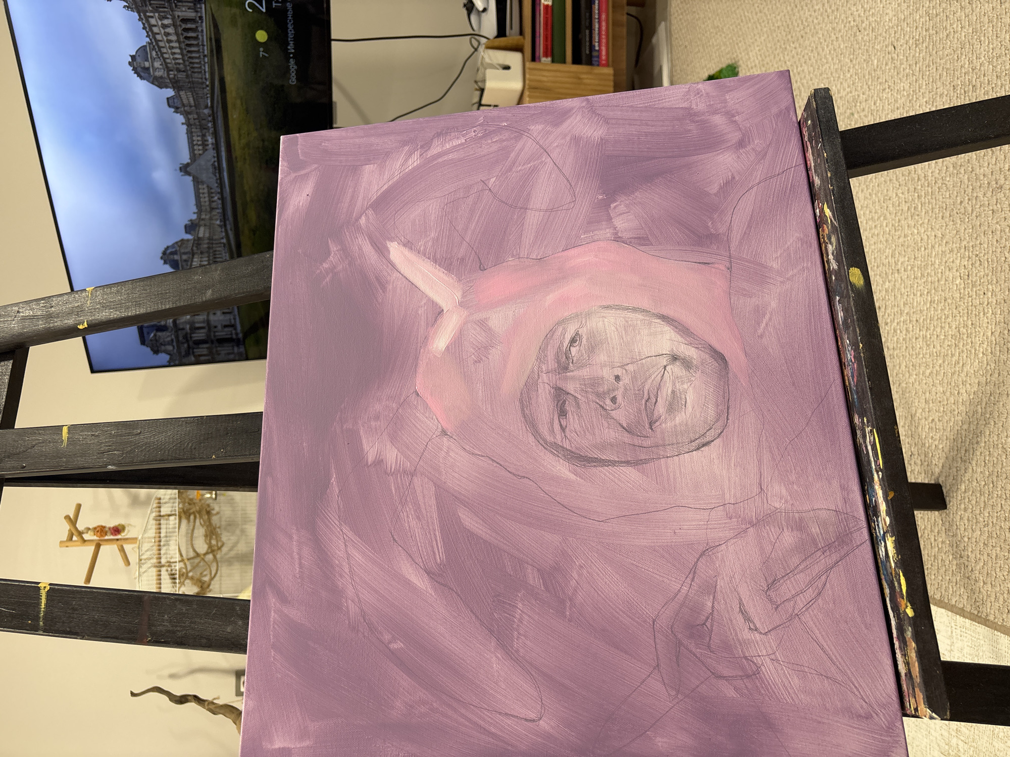 A partially completed portrait painting sits on an easel. The canvas is predominantly covered in light purple paint, with a faint pencil sketch of a face and figure visible. The face appears to be centered within the canvas, surrounded by a softer pink hue. There is a faint outline of a hooded garment on the top of the head. The easel is made of dark wood. Behind the easel, there's a television displaying a building that looks like a European palace, possibly the Louvre in Paris. Adjacent to the TV, there's a bookshelf filled with books and some electronic equipment. A bird cage with a small toy is visible behind the easel. The flooring appears to be carpet. The location appears to be an indoor space in an unidentified private residence. This is not in Krasnoyarsk, Russia.
