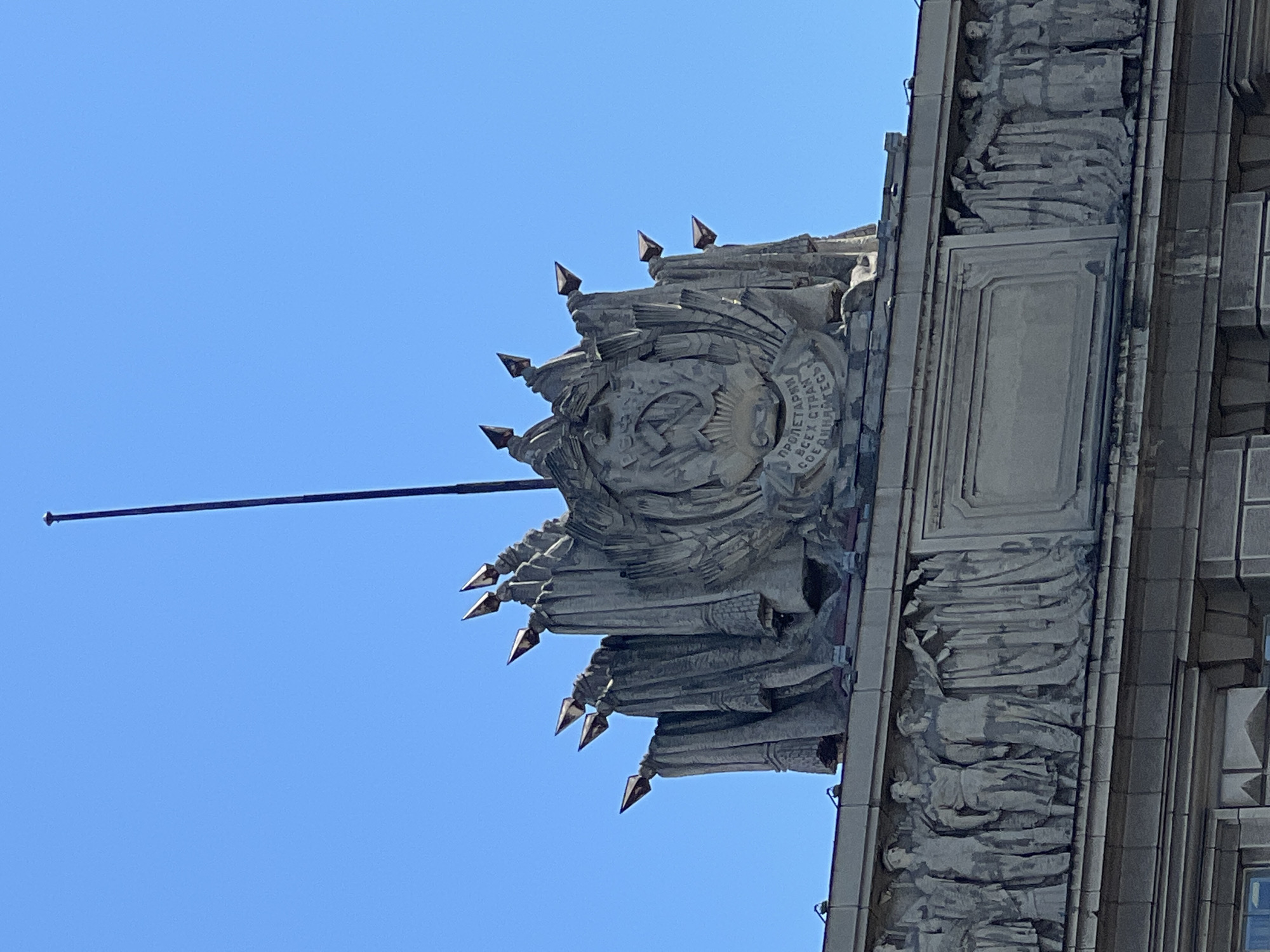 The image shows a detail of the architectural facade of the Leningrad Soviet building in Saint Petersburg, Russia. The upper portion features a prominent sculptural composition against a clear blue sky. This composition is a large, grey stone relief depicting a central Soviet emblem surrounded by stylized figures and symbolic elements. The emblem includes a hammer and sickle. Below the central relief, there is a frieze of smaller, carved figures in relief, representing people in Soviet-era attire, standing in a line and appearing to be engaged in some collective activity. The building itself is constructed of grey stone, and the overall impression is of monumental Soviet architecture.
