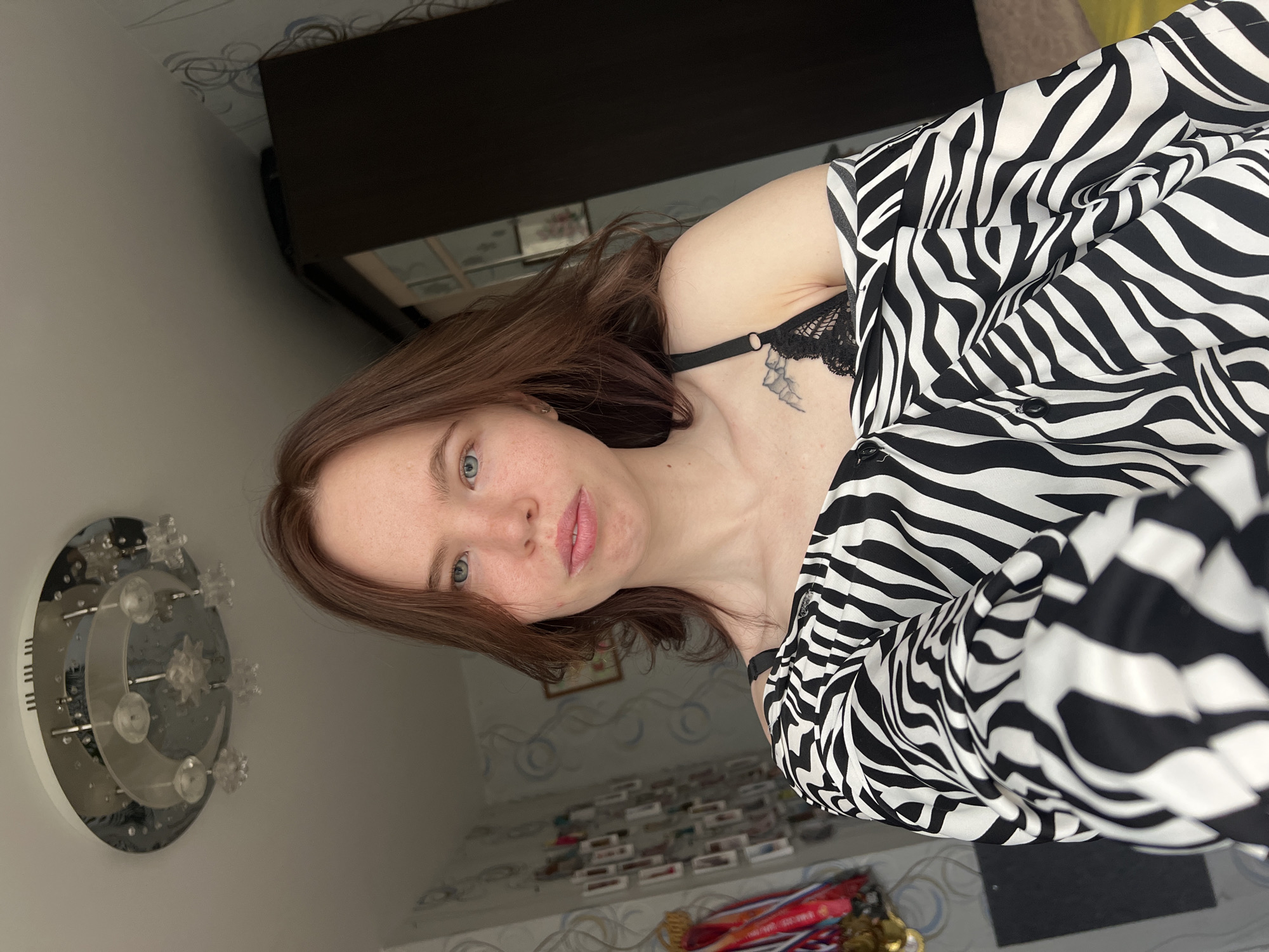 The image displays a young woman posing for a photo in an indoor setting, identified as Chaikovsky, Russia. The photo is rotated 90 degrees clockwise, showing the woman lying on her side, looking directly at the camera. She has light brown hair, striking light blue eyes, and fair skin, with a neutral and composed expression. She is wearing a black and white zebra-print top or robe, with a black lace bralette or bra strap visible underneath. A small, delicate tattoo, possibly depicting a floral or leafy design, is visible on her upper chest/shoulder area.
The background elements suggest a bedroom or personal living space. To the woman's left, a silver-toned, circular wall-mounted light fixture or decoration is visible, featuring clear, star-shaped and crescent moon-shaped accents. Behind her head, there is a dark wooden piece of furniture, possibly a dresser or cabinet, with a subtle patterned wallpaper on the wall above it. In the lower part of the frame, below her, several small, rectangular items (possibly cosmetic products or boxes) are neatly arranged on shelves or a surface, accompanied by a collection of medals displayed on ribbons. The overall lighting suggests it is daytime. No visible text is present in the image.