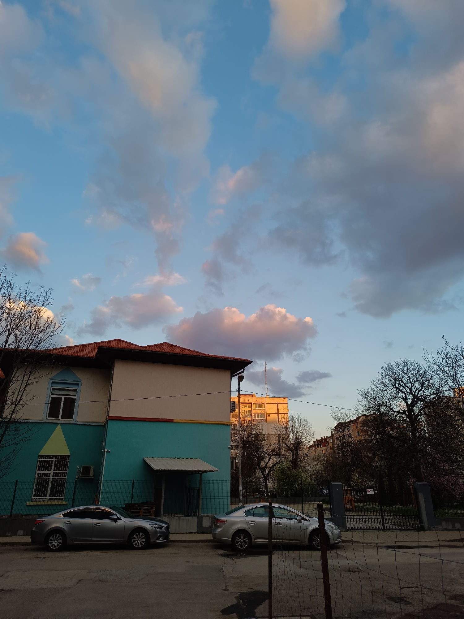 The image shows a street scene in what appears to be a residential area. Two light grey Mazda 6 cars are parked on the side of the street in front of a building. The building is two-story with a red tile roof. The upper part of the building has white walls with a tan stripe while the lower part is painted in a turquoise blue color. A green triangle design is present on the wall to the left of the image. Across the street, there is a black metal fence and a park with trees. Several buildings of 5 to 7 stories can be seen in the distance. The sky is partly cloudy with white and grey clouds. Some clouds have a pink hue from the setting sun. The location is most likely Sofia, Bulgaria.
