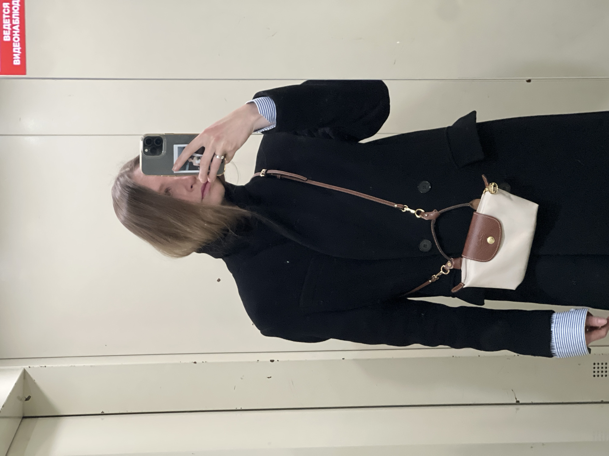 A person stands in front of a mirror, taking a selfie with a smartphone. The individual is wearing a black coat, a turtleneck, and a blue striped shirt peeking out from under the coat sleeves. They have light brown hair. A small off-white bag with a brown leather flap and strap hangs from their shoulder. The mirror reflects a pale wall behind the person. In the upper left corner of the mirror, a red sign with white text, apparently in Russian, is visible. The photograph was taken in Khimki, Russia.