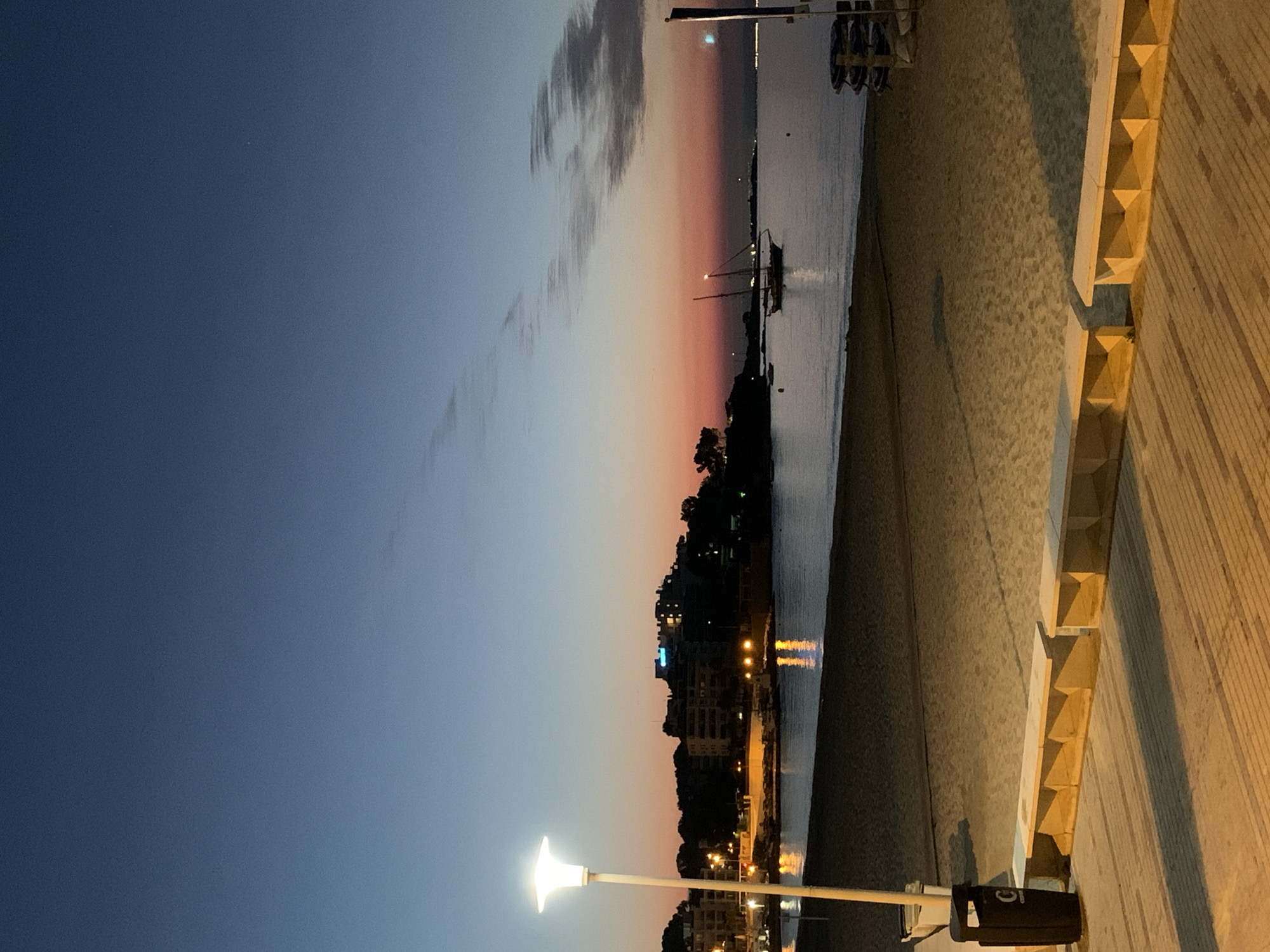 A wide shot captures a beach in Palmanova, Spain, at dusk. The sky transitions from a deep blue at the top to a soft orange and pink near the horizon, with faint clouds streaking across. A lone, illuminated lamppost stands on the left side of the frame, casting a bright glow. In the foreground, a wooden promenade is lined with beige, geometric benches. A black trash receptacle is positioned next to the lamppost. The sandy beach stretches out towards the water, where the gentle waves lap the shore. The sea reflects the lights from a distant town, which features several buildings illuminated in the twilight. A sailboat with two masts is anchored in the water, adding a touch of nautical charm to the scene. The overall atmosphere is serene and tranquil, suggesting the end of a day at the beach.