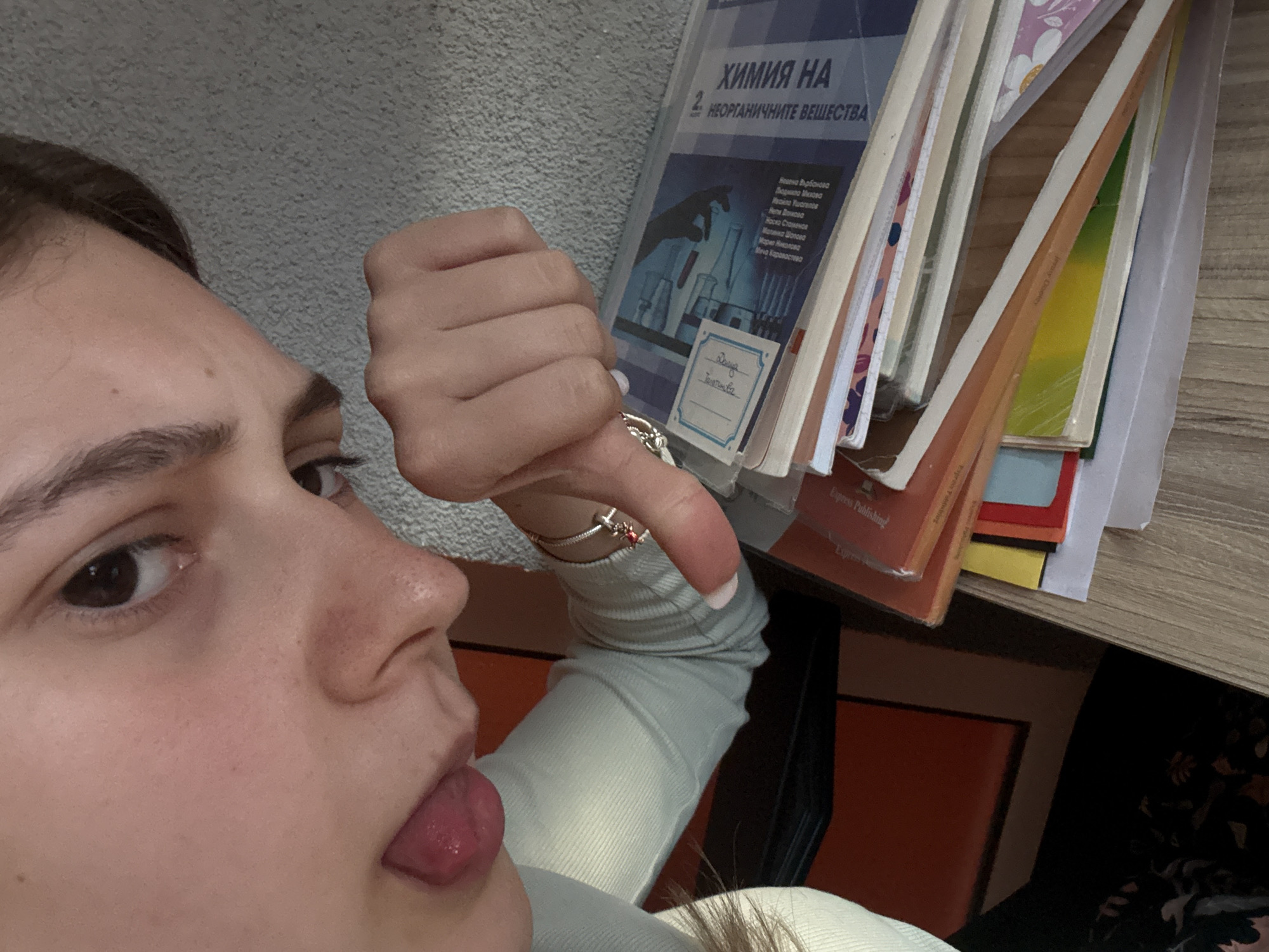 A young person, likely female, is shown in a close-up, diagonally oriented shot, playfully making a "thumbs down" gesture with one hand and sticking out their tongue. The individual wears a light-colored, long-sleeved top and accessorizes with a ring on their finger and a bracelet on their wrist.
Behind the person, a stack of academic books rests on a light-colored wooden shelf, with a grey, textured wall forming the backdrop. The most prominent book is titled "ХИМИЯ НА 2. НЕОРГАНИЧНИТЕ ВЕЩЕСТВА" (CHEMISTRY OF INORGANIC SUBSTANCES). Authors listed include Невена Върбанова, Людмила Михова, Ивайла Ушагелов, Неля Донкова, Наско Стаменов, Малинка Шопова, Мария Николова, and Меча Каравастева. A handwritten label on this book displays "Даяна" and "Тодорива," likely a student's name. Other visible text on book spines includes "Jenny Cloetey" and "Express Publishing." The setting appears to be an indoor study or academic environment. The activity suggests a playful reaction, possibly expressing disinterest or mild disapproval towards the subject matter of the books. This scene is situated in Panagyurishte, Bulgaria.