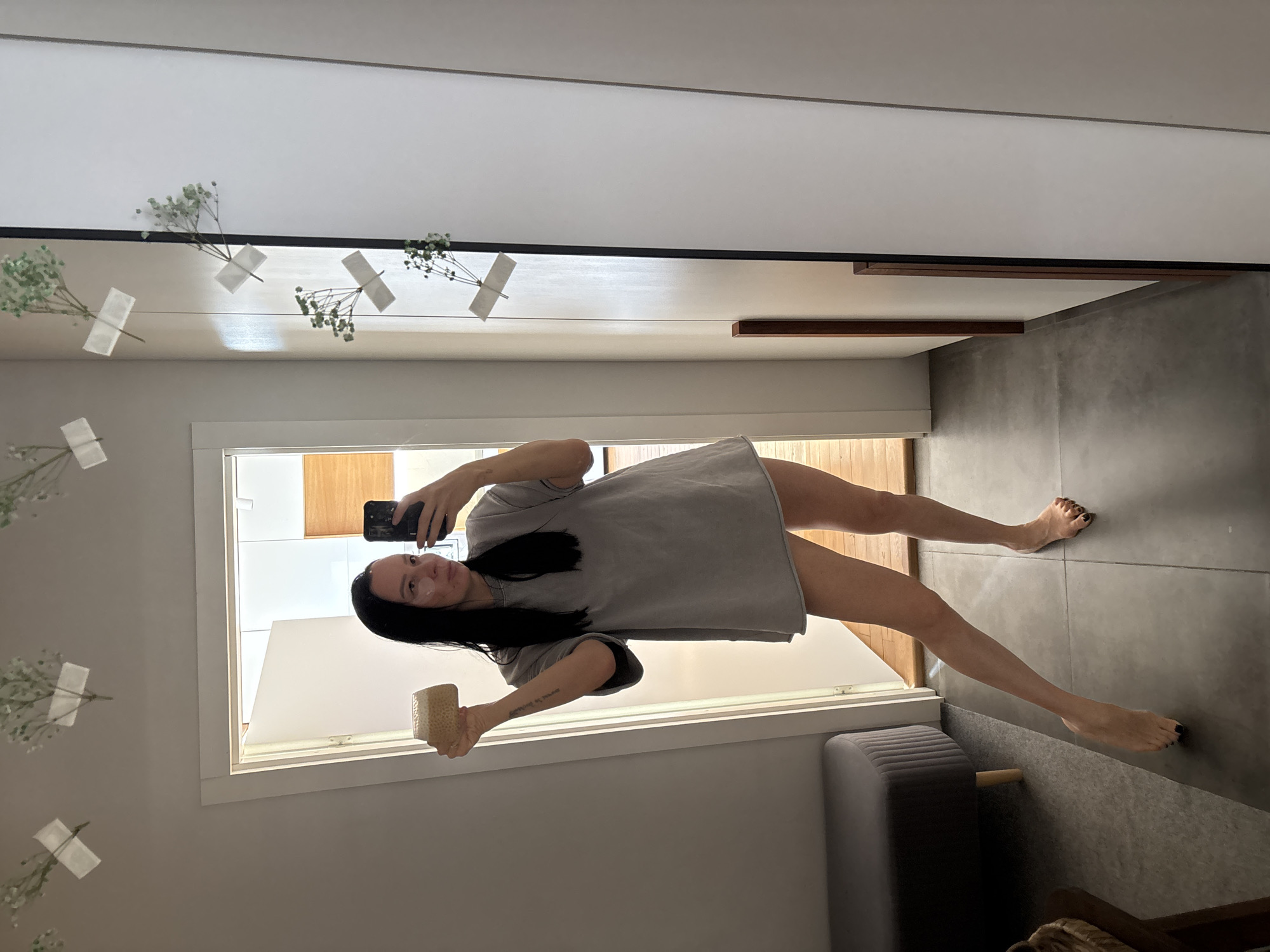 A young woman stands indoors, taking a mirror selfie with her smartphone. She holds the phone in her right hand to capture her full reflection, while her left hand holds a light-colored, textured object, possibly a natural sponge or body brush. She has long, dark hair and is wearing a loose-fitting, light gray t-shirt or short dress. A small, light-colored facial patch is visible below her left eye. A tattoo on her left forearm reads "Dreams of you." Her bare legs extend towards the floor, and her toenails are painted a dark color.
The setting is a modern indoor space, likely a dressing area or bathroom, in Chaikovsky, Russia. The room features light-colored walls and ceiling, complemented by dark gray tiled flooring. A large window or open doorway in the background allows natural light to illuminate the scene, indicating it is daytime. Horizontal wooden accents are integrated into the wall or ceiling design. Delicate sprigs of baby's breath, or similar small white flowers, are taped to the upper wall or ceiling, forming a decorative garland. A gray upholstered bench or ottoman is visible in the lower right, along with a partial view of a wicker item, possibly a basket. The woman has a slight smile, suggesting a relaxed or happy mood.