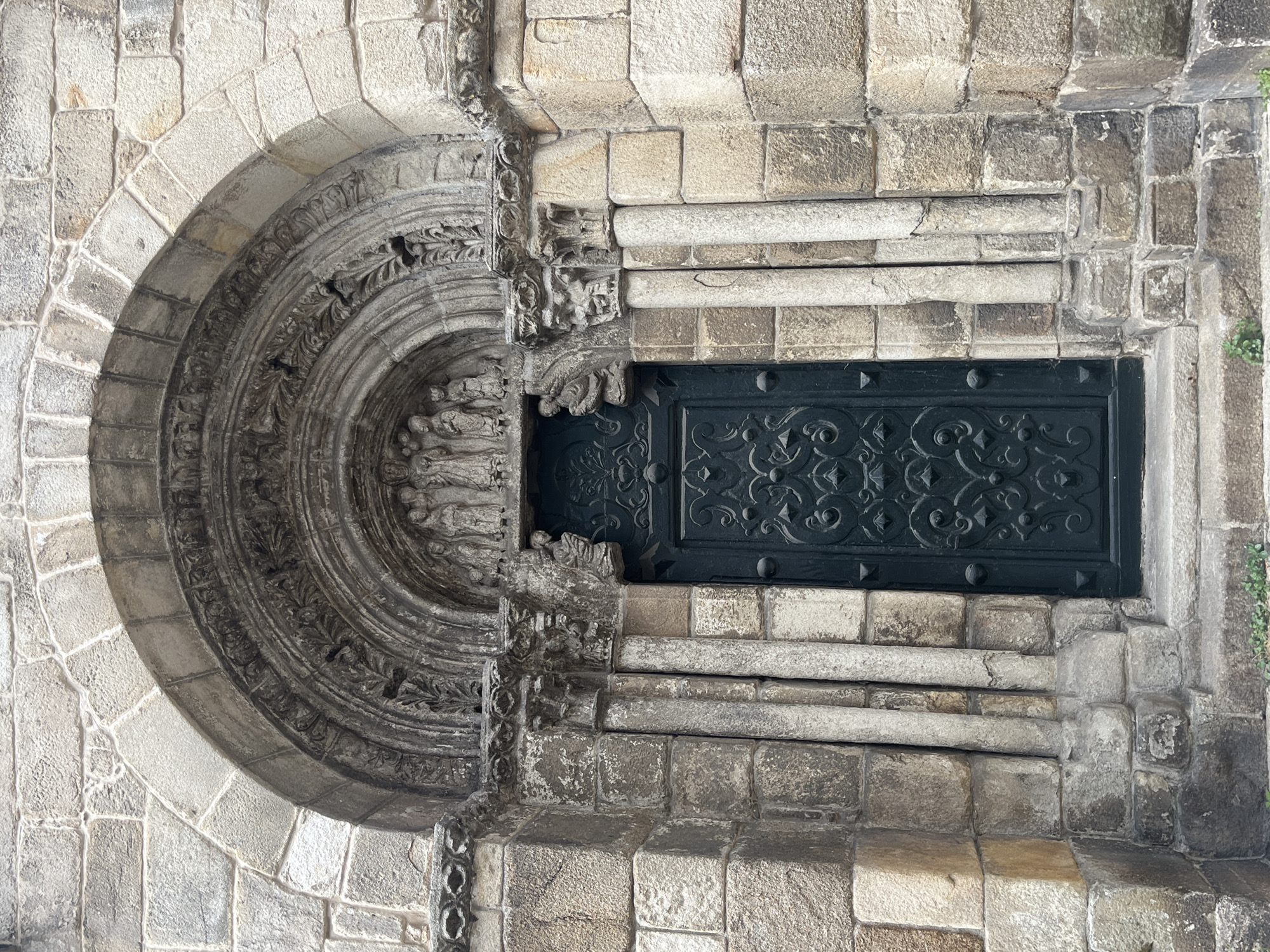 The image shows a detailed stone entrance with a semicircular arch, heavily ornamented with carved details. The doorway is partially blocked by a dark, intricately patterned door. The stone is weathered, suggesting age. The scene depicts the Romanesque portal of the Church of Santiago in A Coruña, Spain. There are no people visible.