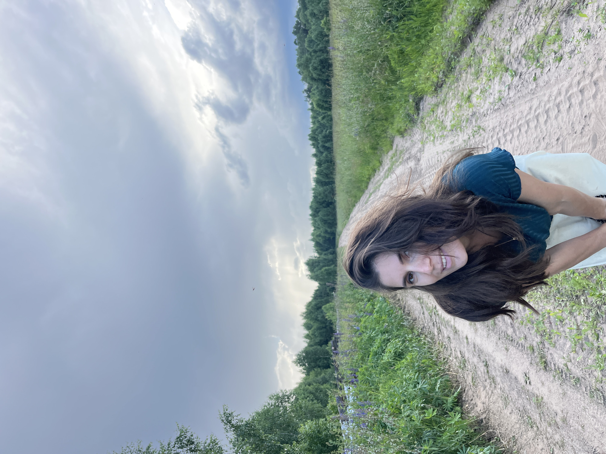 The media file is a portrait of a young woman with long, dark brown hair, taken outdoors in a rural setting. She is looking towards the camera with a slight smile and her tongue peeking out. She is wearing a dark green top and a light-colored garment, possibly a skirt or dress.
The scene appears to be a sandy path leading through a field of green grass and wildflowers, with a dense line of trees in the background. The sky is cloudy, with patches of sunlight breaking through, suggesting either early morning or late afternoon. The overall mood is casual and relaxed, with the woman's pose and expression conveying a sense of playfulness.
The location is identified as Уральское, Russia, which provides context for the natural landscape. There is no visible text in the image.